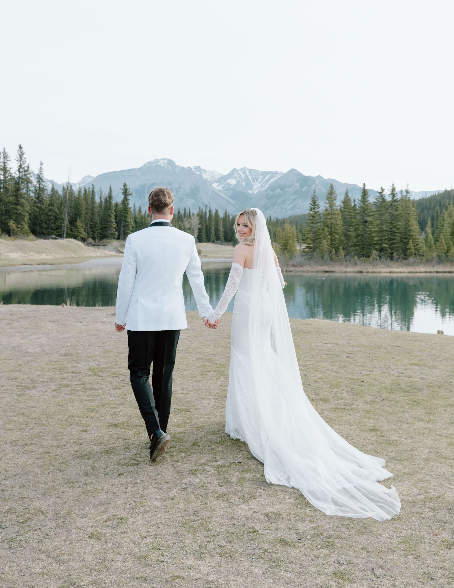 Couple holding hands walking by mountain lake in wedding attire. Calgary Wedding Photographer