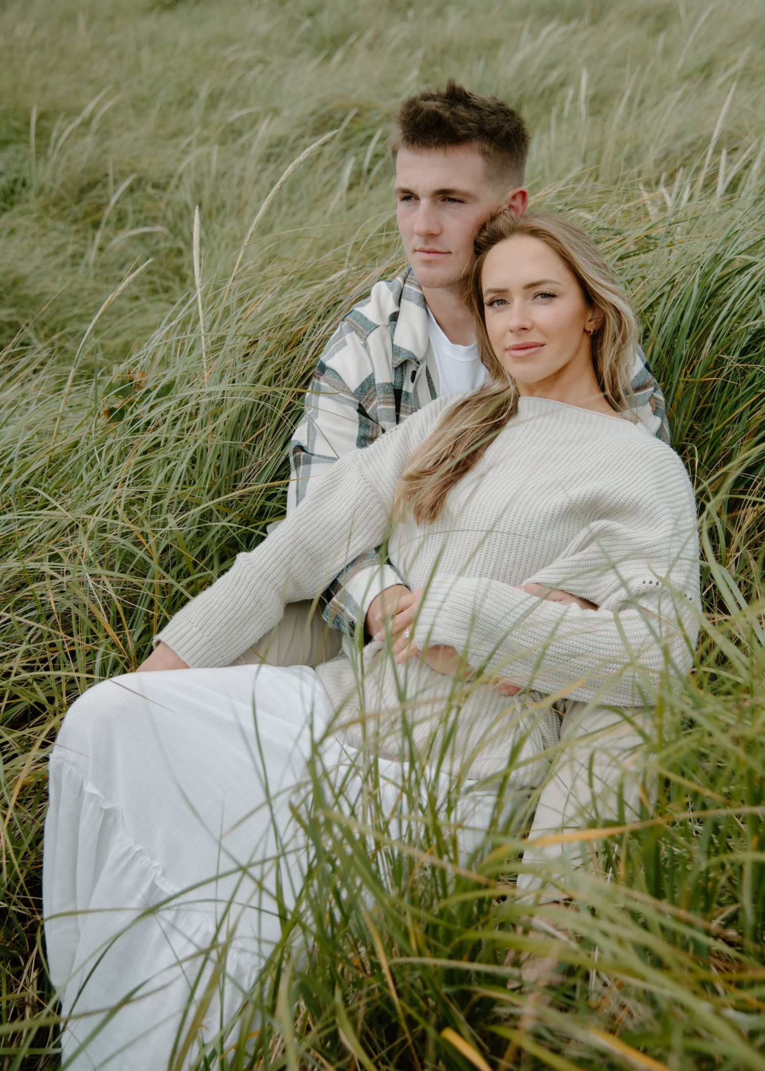 Romantic couple lounging in tall grass on coastal dunes wearing coordinated neutral outfits. Calgary Alberta Engagement Photographer.