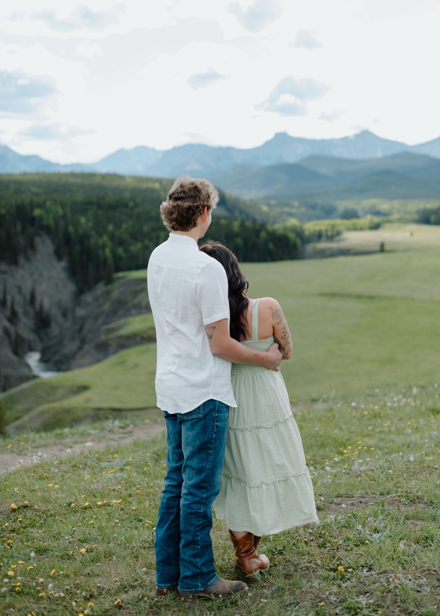 Two people embrace while taking in panoramic mountain views on a grassy hillside in Calgary, Alberta Calgary Lifestyle and Engagement Photo
