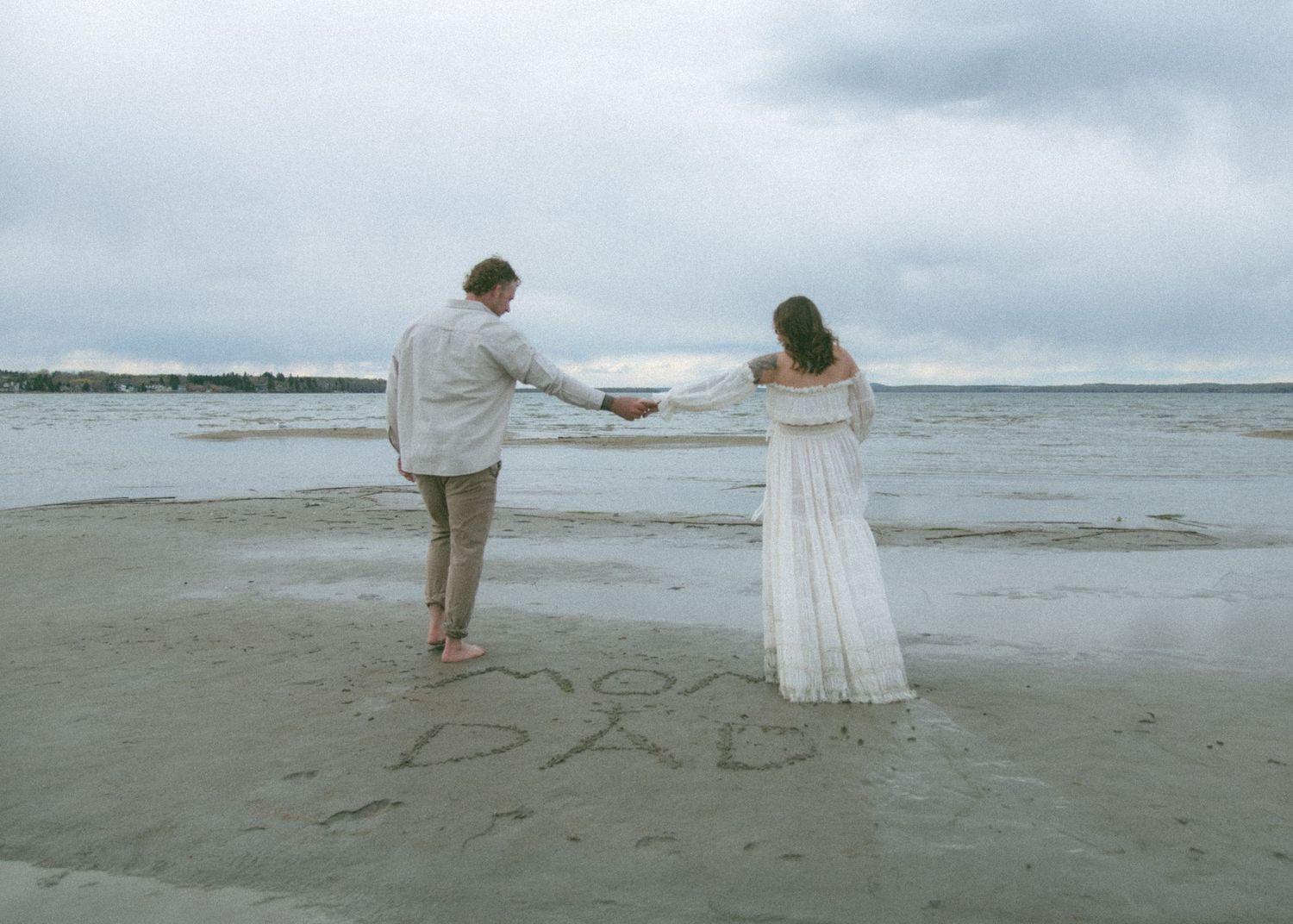 A couple in white attire holding hands while walking on a beach with text written in sand. Calgary Alberta Maternity Photographer.