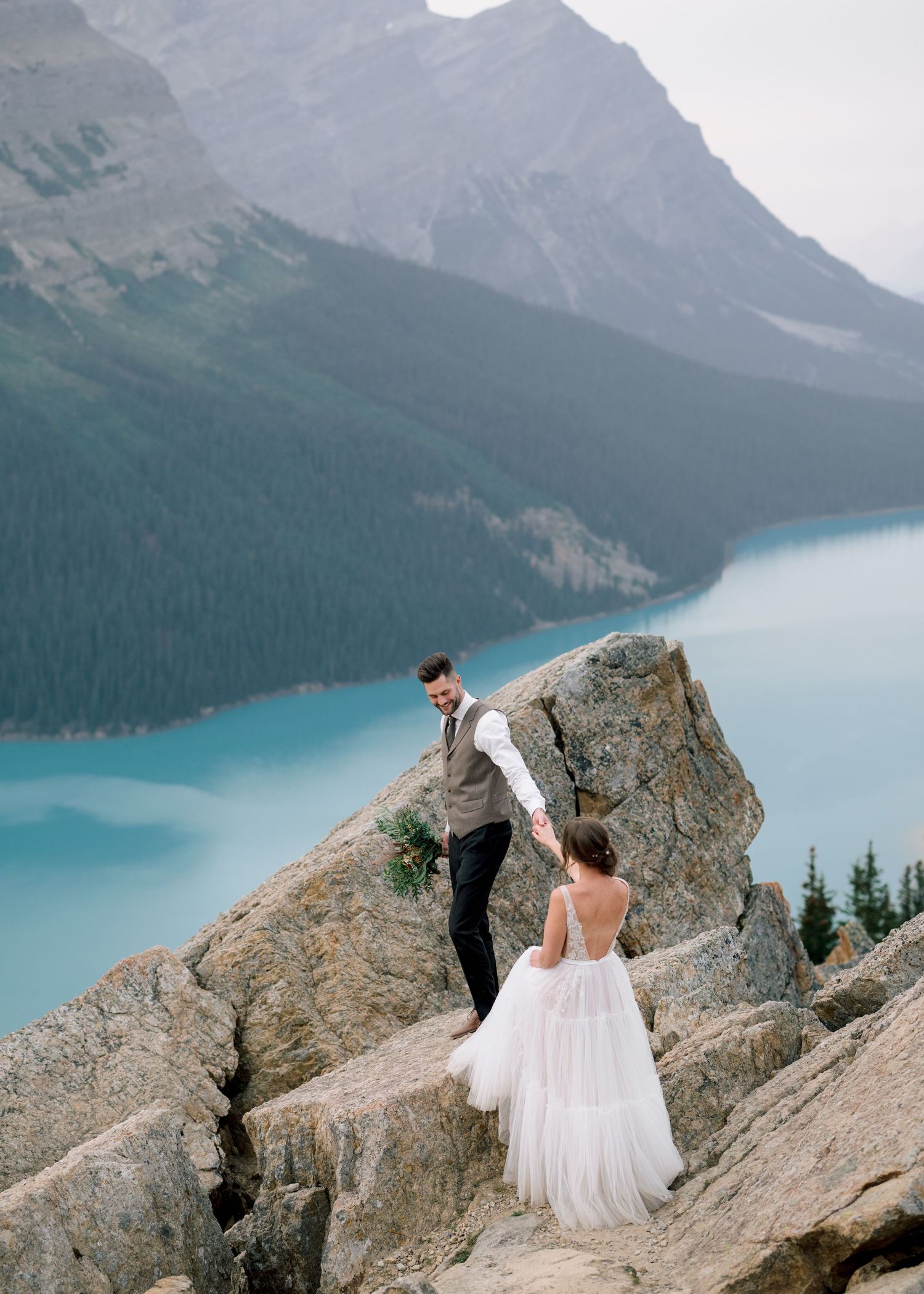 Two figures in formal attire stand on rocky cliffs overlooking turquoise glacial lake and mountain peaks in Banff National Park.