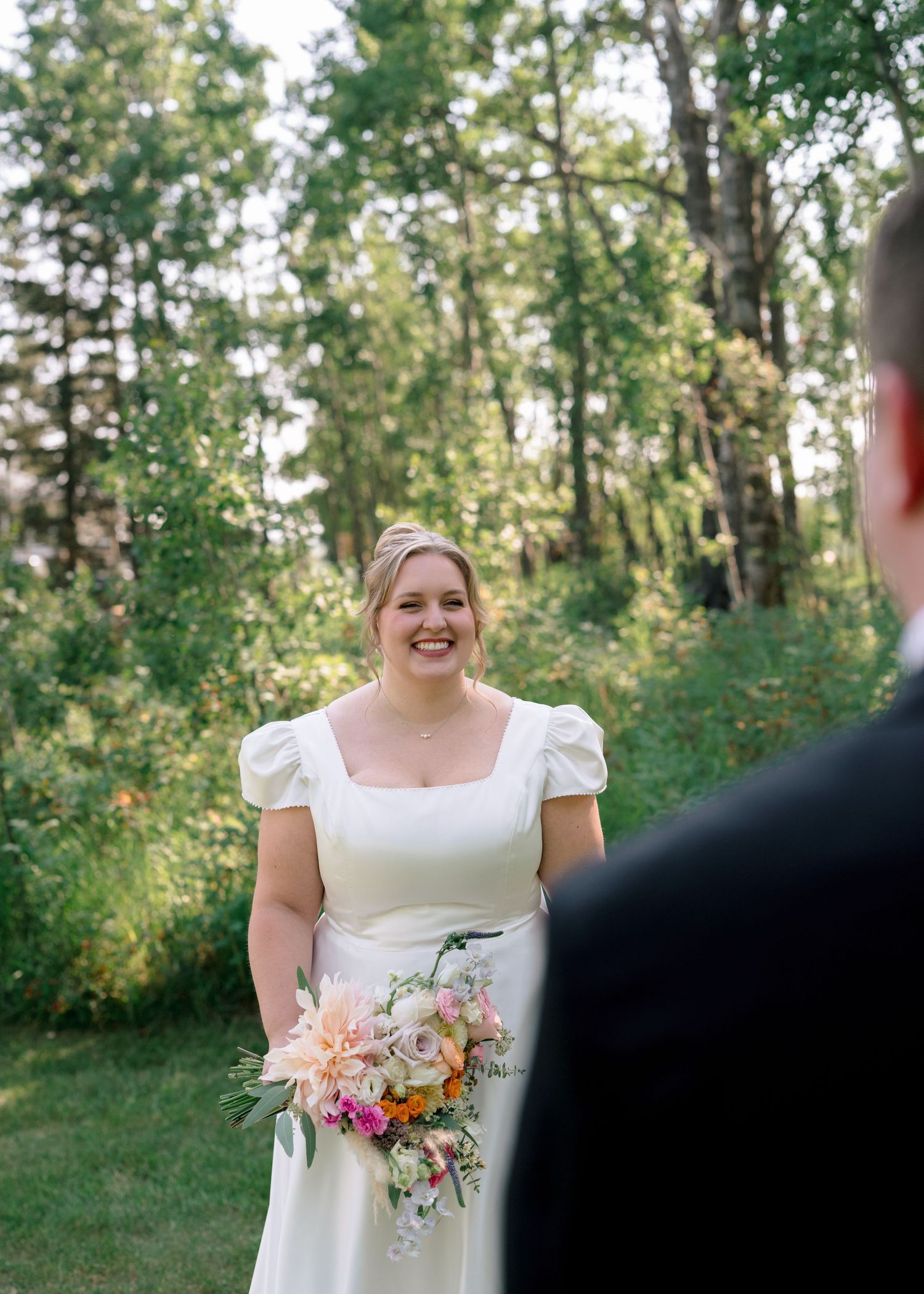 A bride joyfully laughs outdoors while holding a vibrant pink and peach floral bouquet in a natural garden setting.