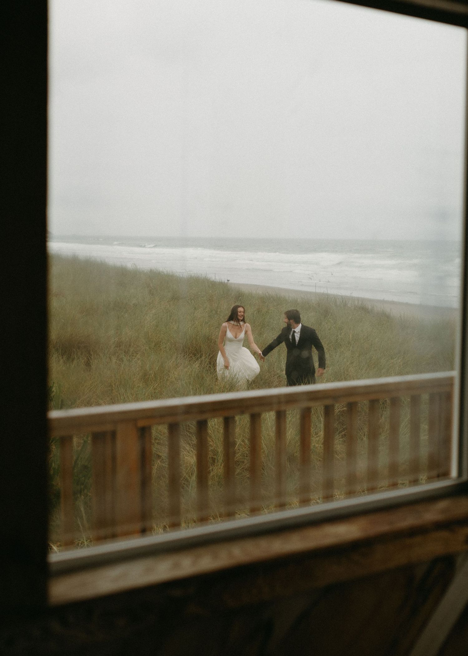 Couple walking together on a wooden boardwalk overlooking a misty beach with tall grass and waves in the distance.
