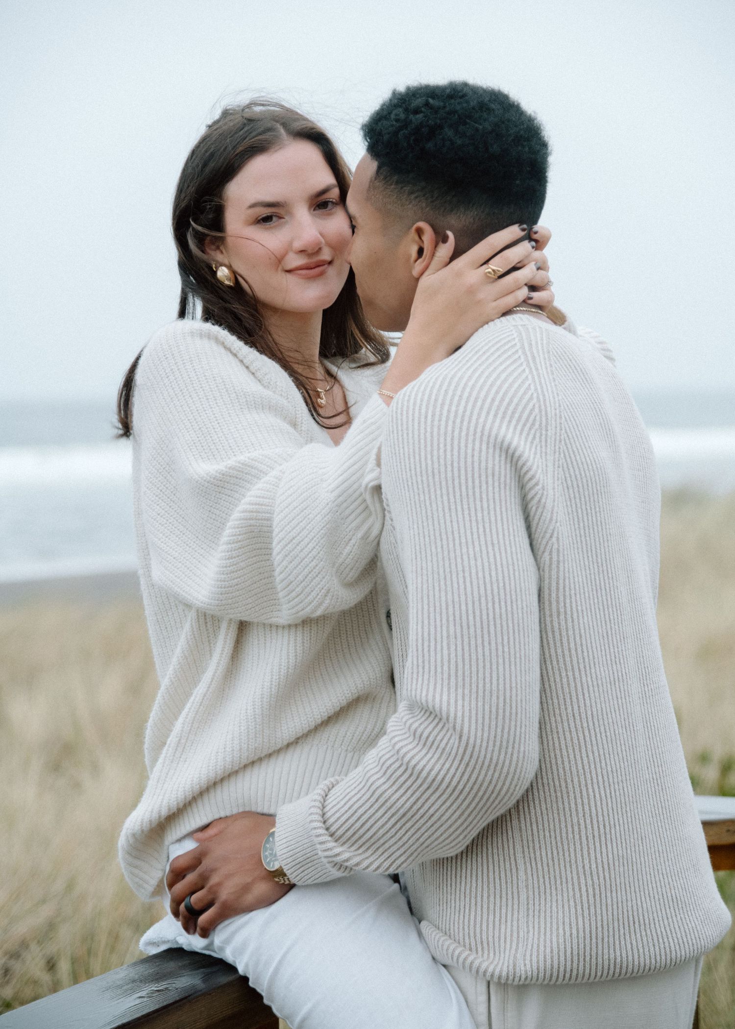 A couple wearing matching cream-colored sweaters embrace lovingly in a scenic outdoor setting by the coast.