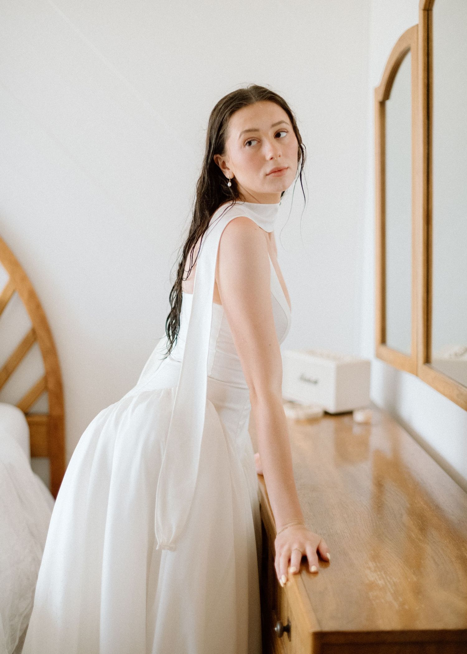 A person in a white flowing dress leans against a wooden desk in a minimalist room with natural light.