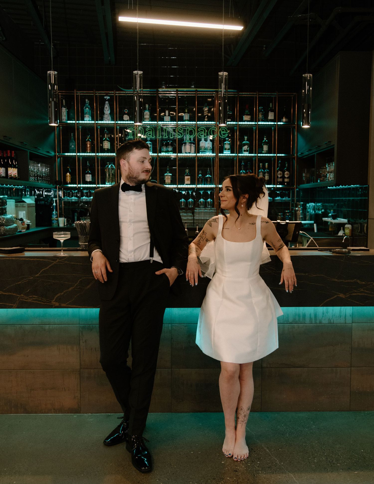 A couple shares a casual moment at a modern bar with teal ambient lighting. Calgary Wedding Photographer