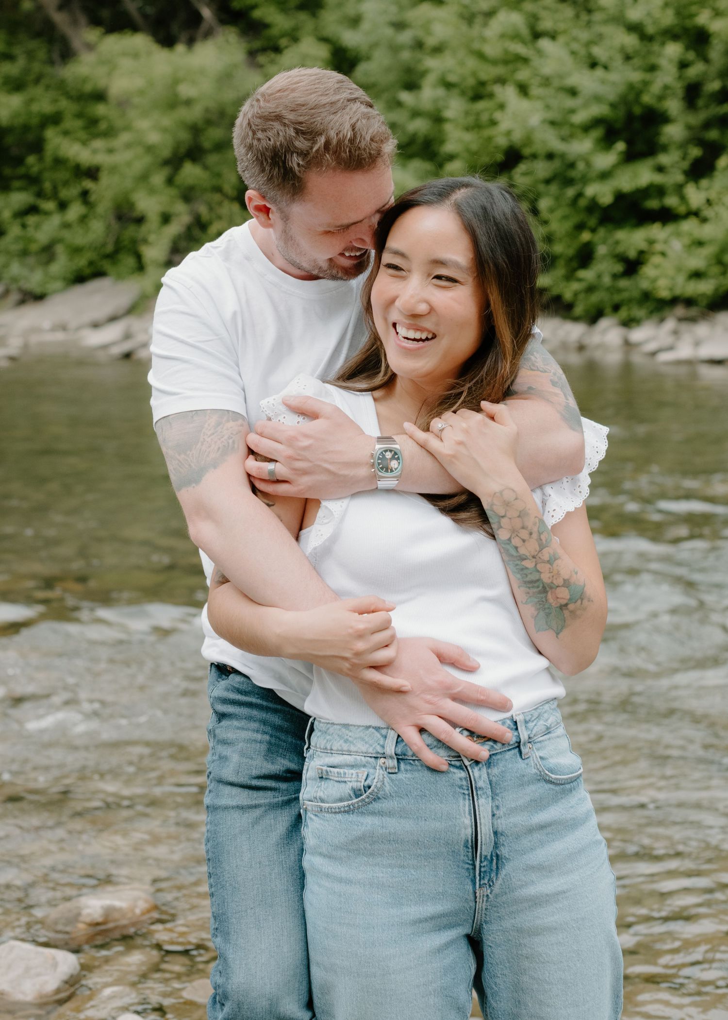A couple shares a tender embrace while laughing together by a flowing river. Calgary Alberta Family and Maternity Photographer.