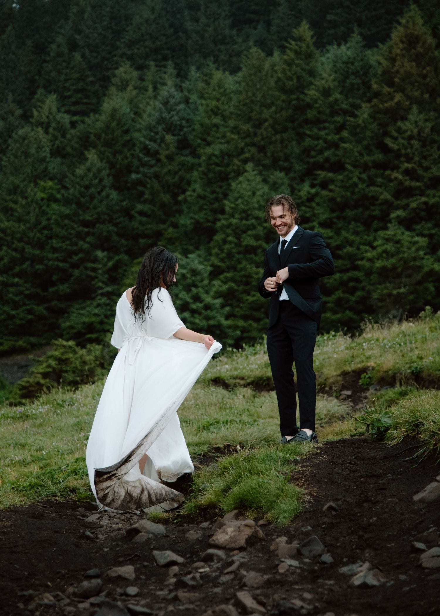 A couple in wedding attire share a joyful moment on a mountainside path surrounded by evergreen trees Banff and Calgary Wedding Photographer