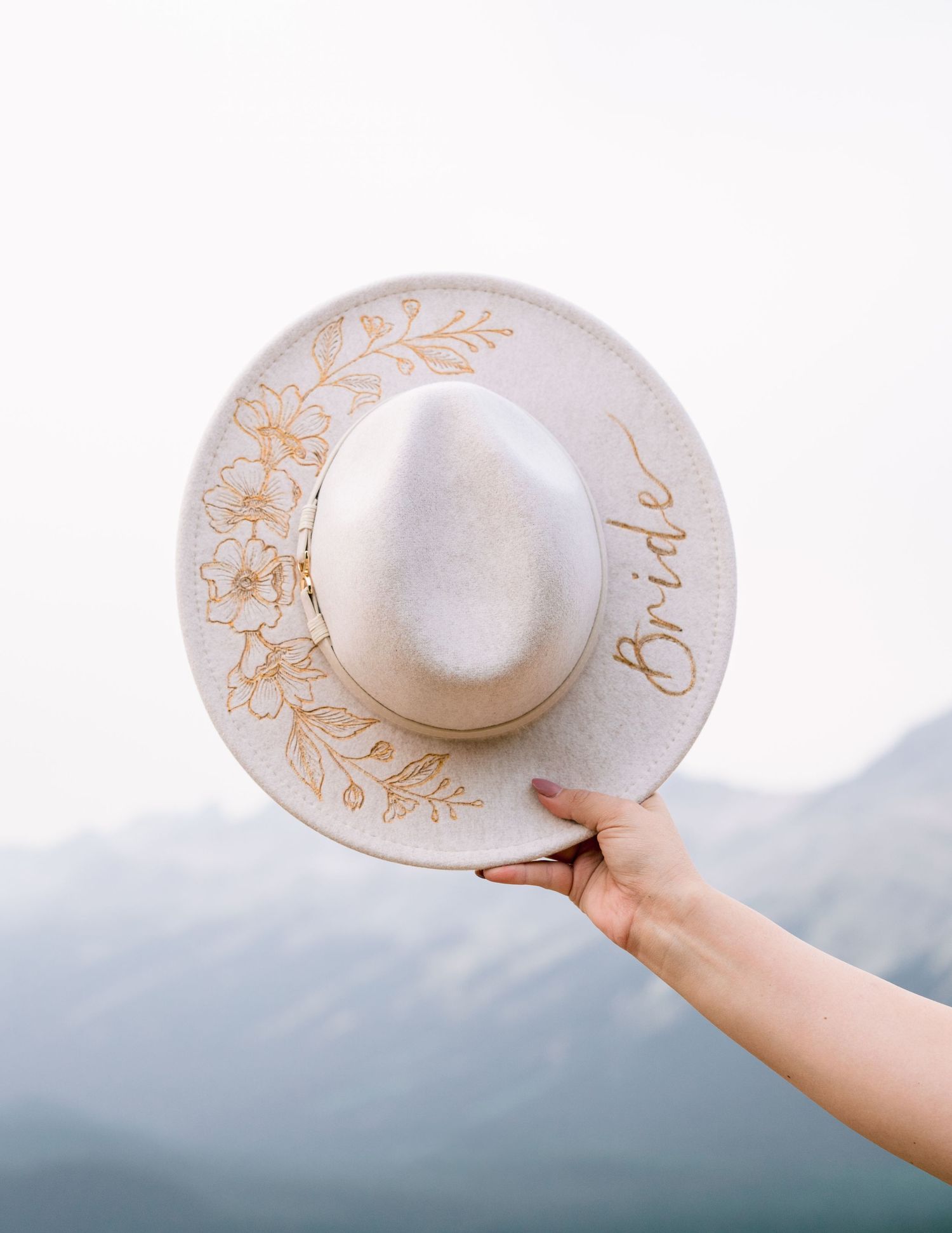White sunhat with gold embroidery held against blurry mountain landscape in dreamy minimalist style.