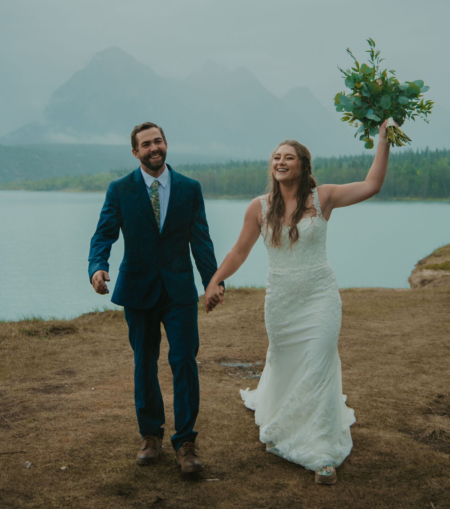 Celebratory wedding couple walking along lakeside trail with mountain vista behind them. Calgary Wedding Photographer