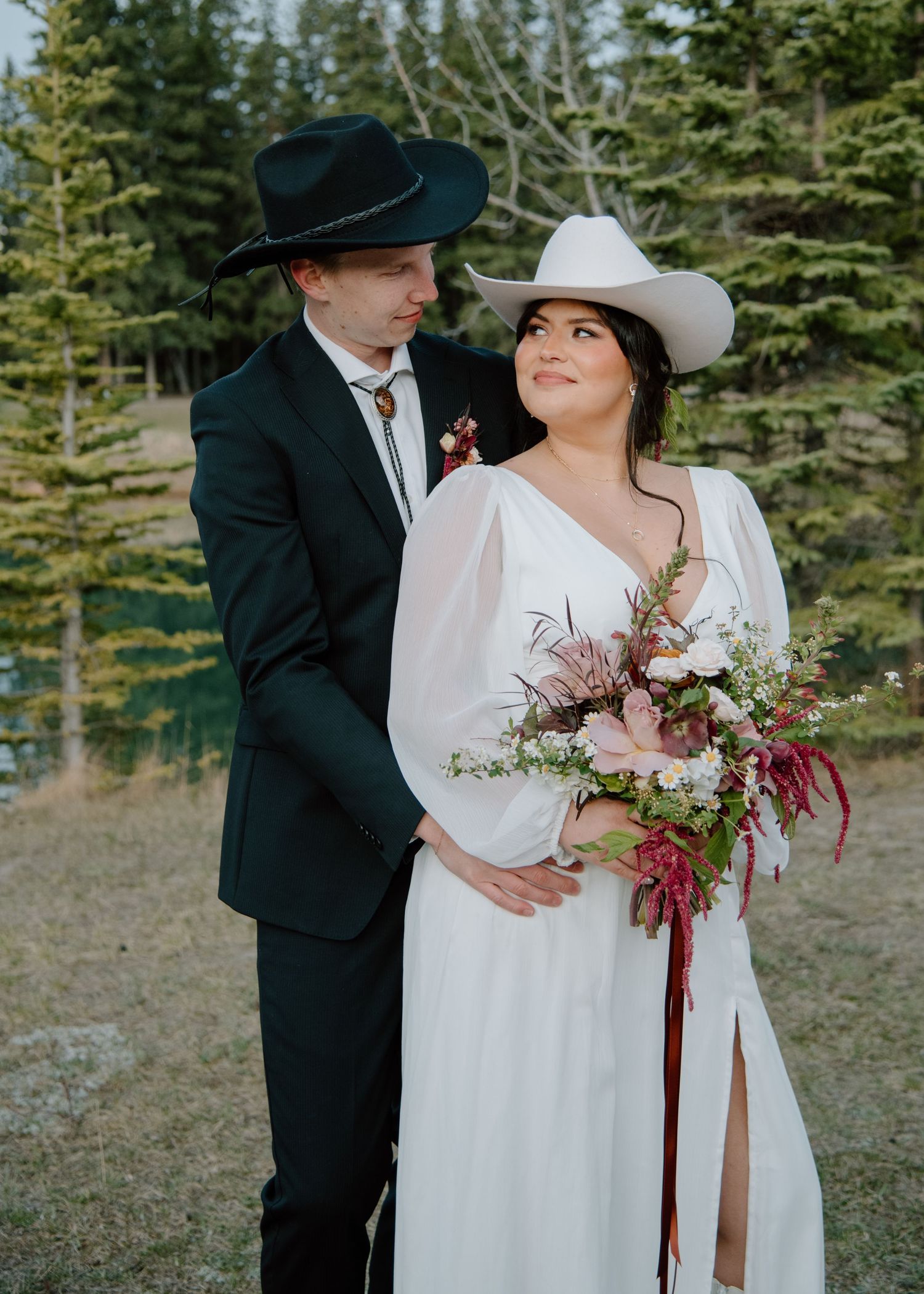 A western-themed wedding couple poses with a beautiful wildflower bouquet by a lake. Banff and Calgary Wedding Photographer