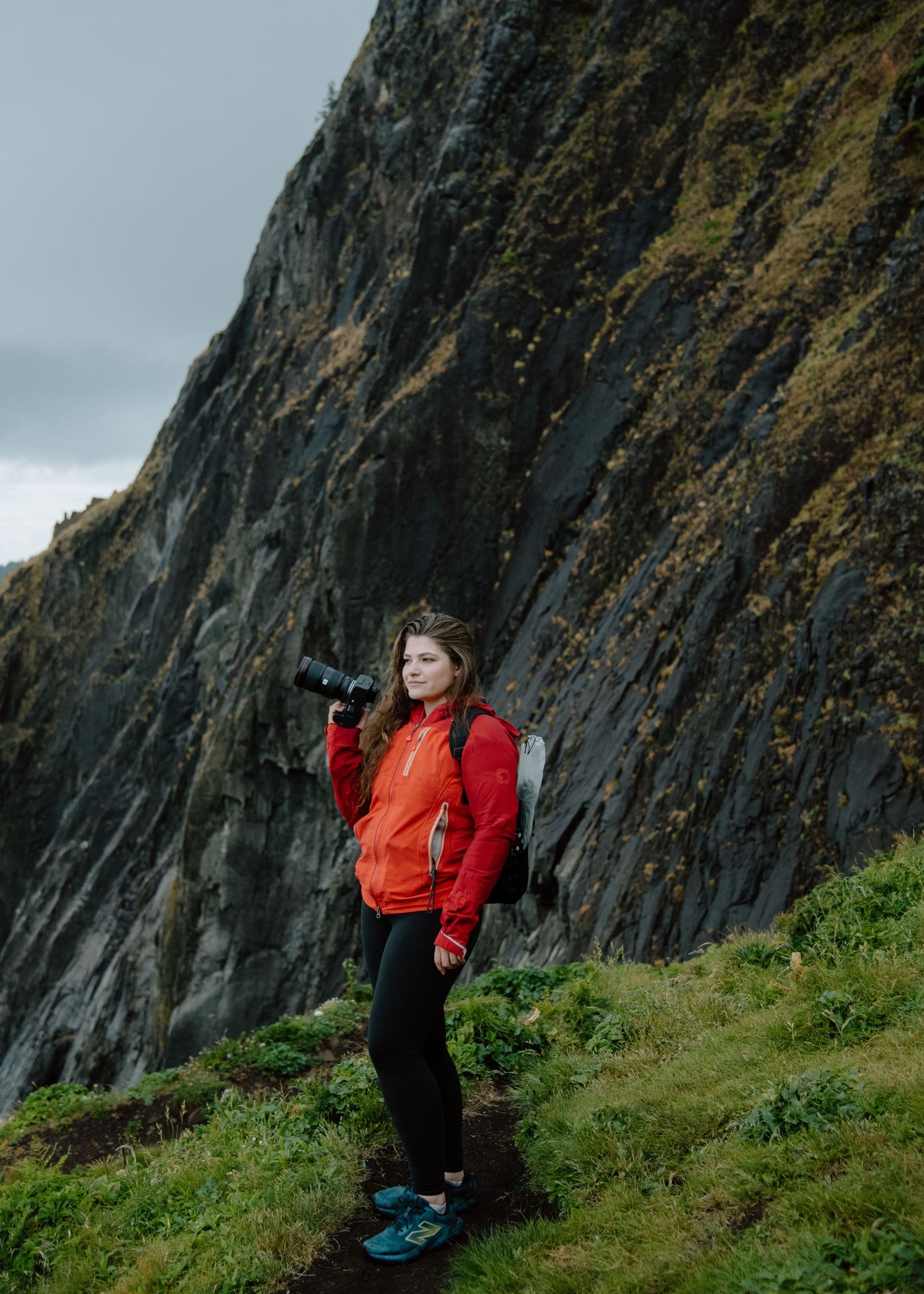 Hiker in red jacket stands on cliff edge with camera against dramatic coastal scenery. Calgary Wedding Photographer