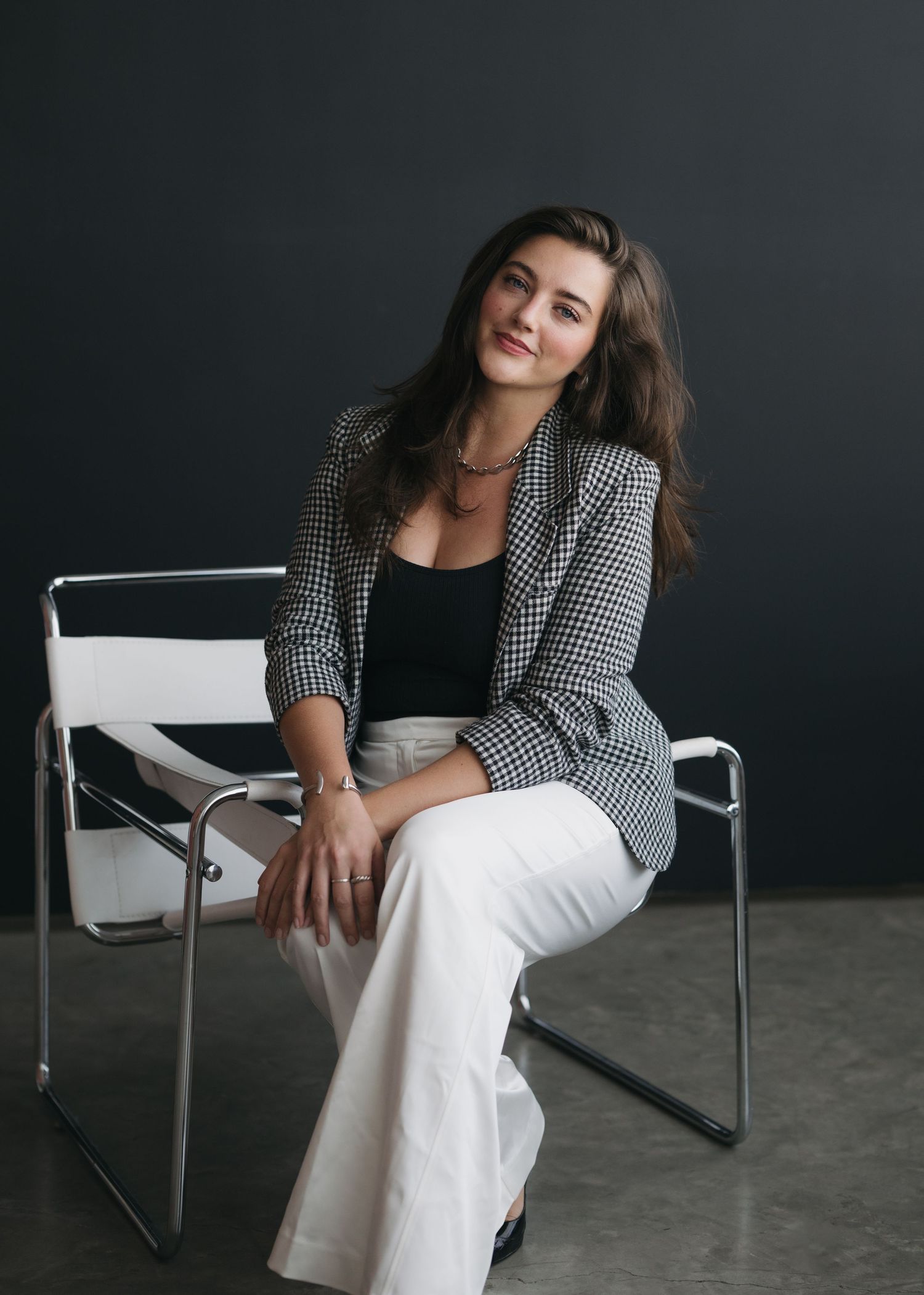 Professional fashion portrait of someone in a houndstooth blazer and white pants sitting casually on a modern white chair.