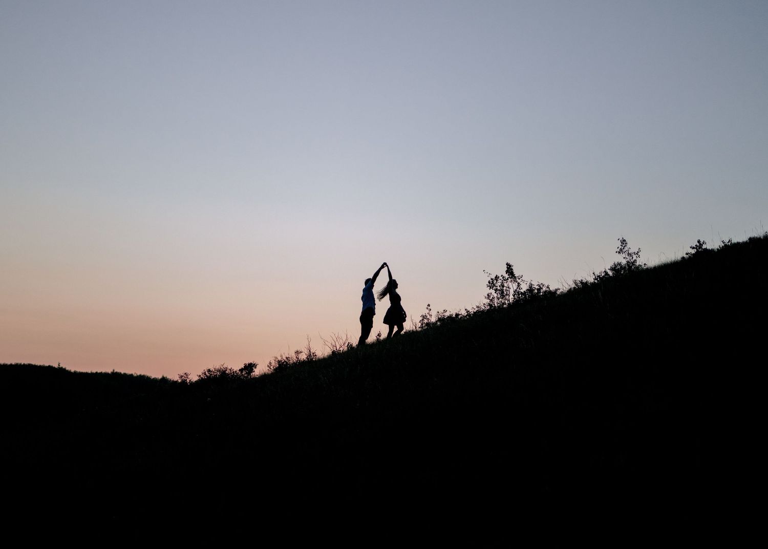 Silhouetted figures stand on a hilltop against a dusky sky at sunset creating a romantic outdoor atmosphere.