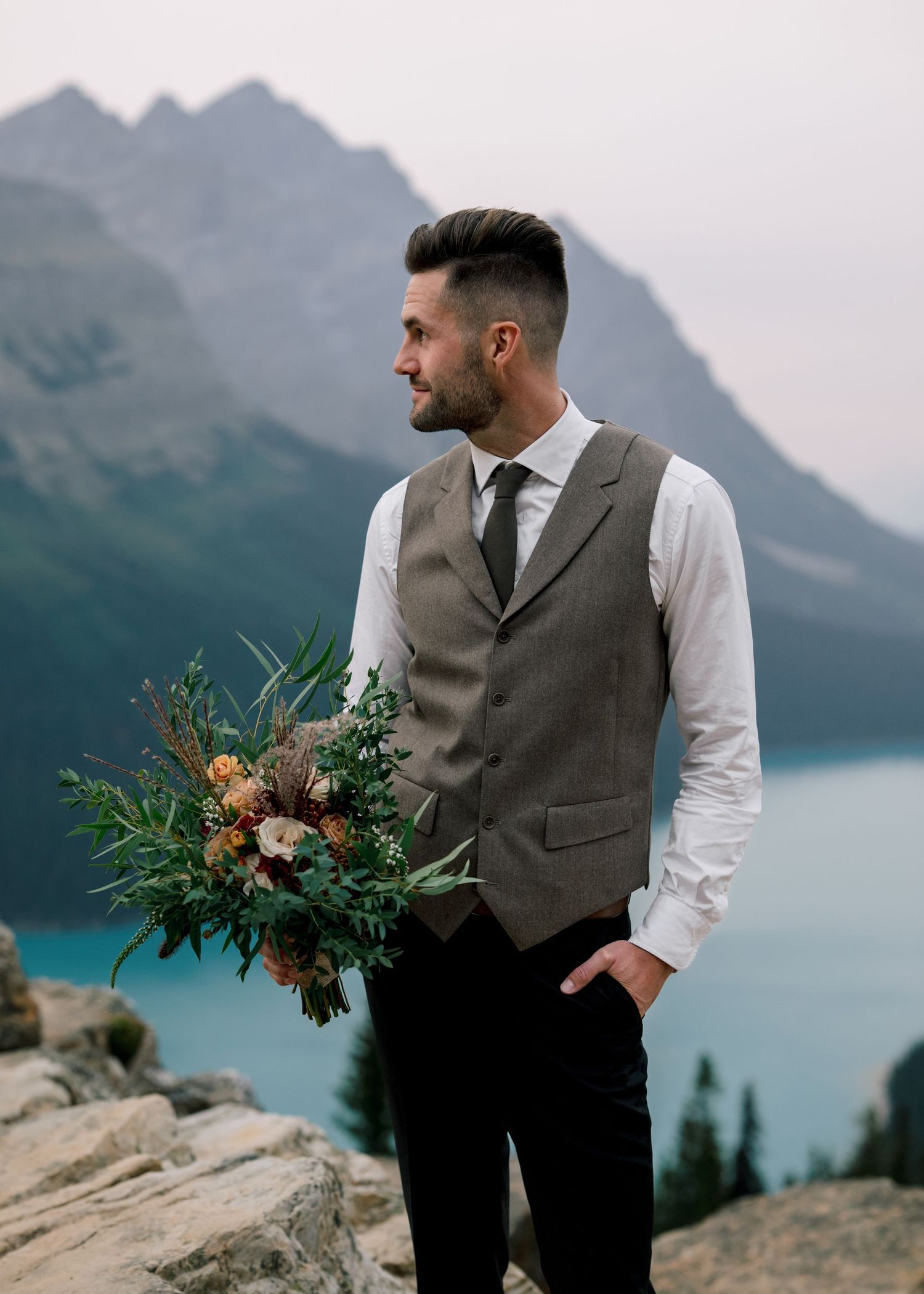 Groom in vest and dress shirt admires scenic mountain lake views at Moraine Lake during outdoor wedding photography session.