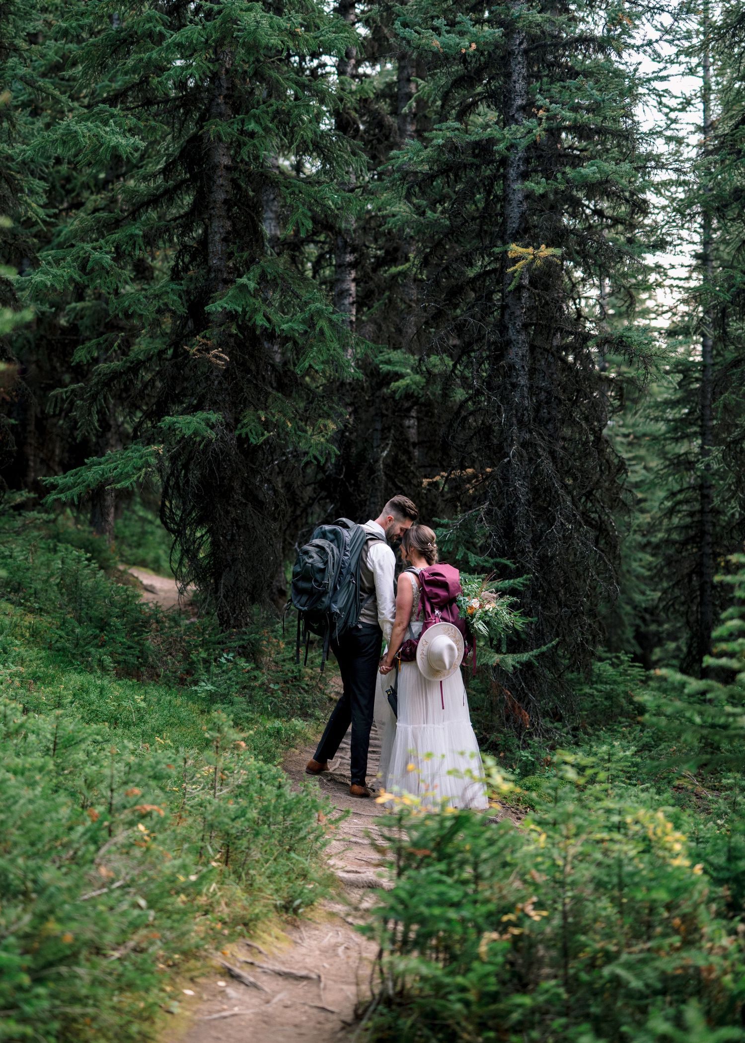 Two figures stand together on a forest trail surrounded by towering evergreen trees and lush greenery on a misty day.