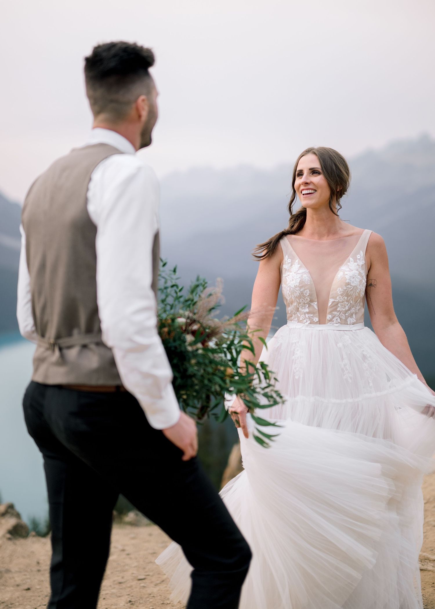 Wedding couple dances and twirls on a scenic mountain viewpoint overlooking majestic peaks.