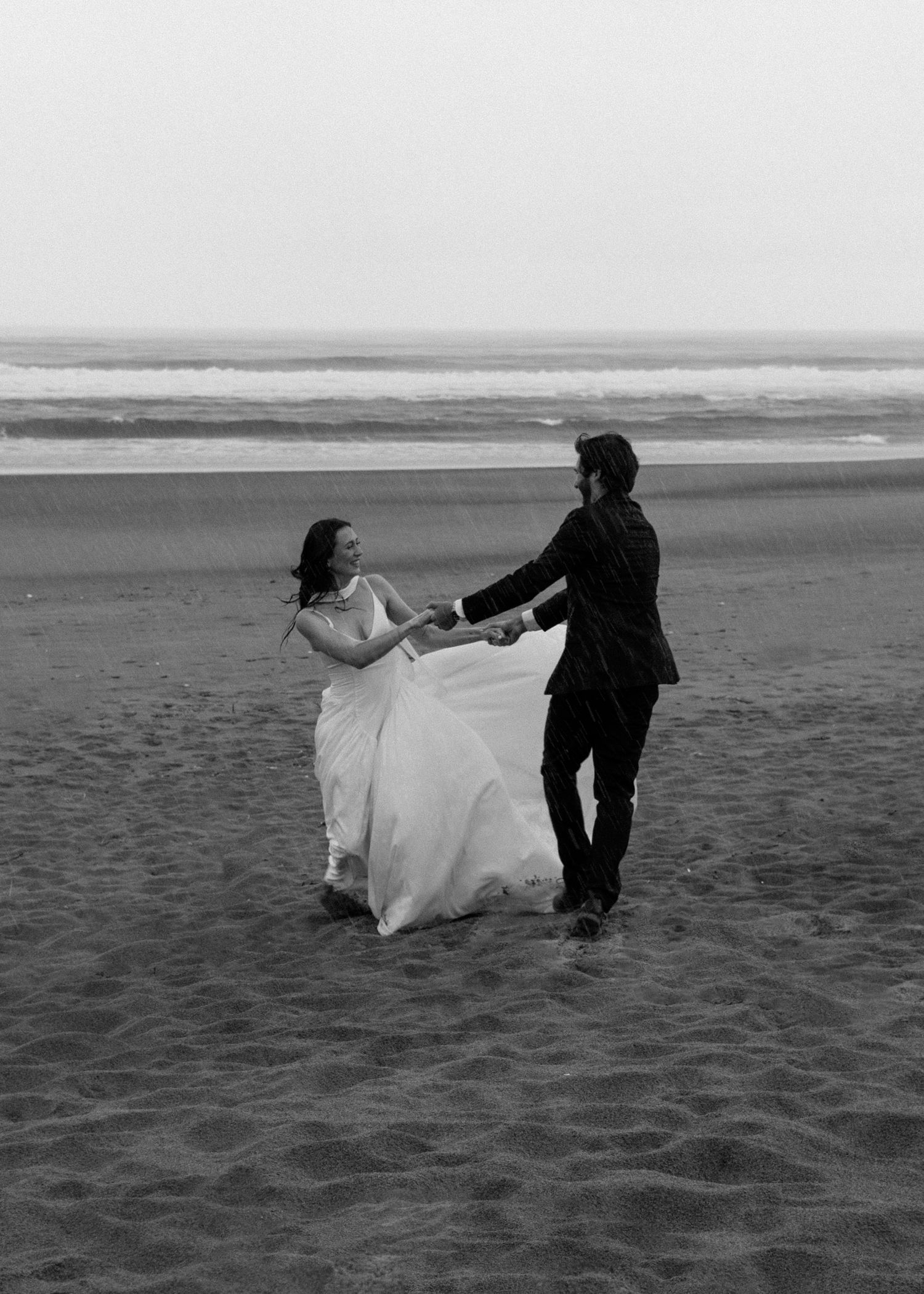 A couple dances romantically on a misty beach in black and white, with the bride's white dress flowing in the wind.