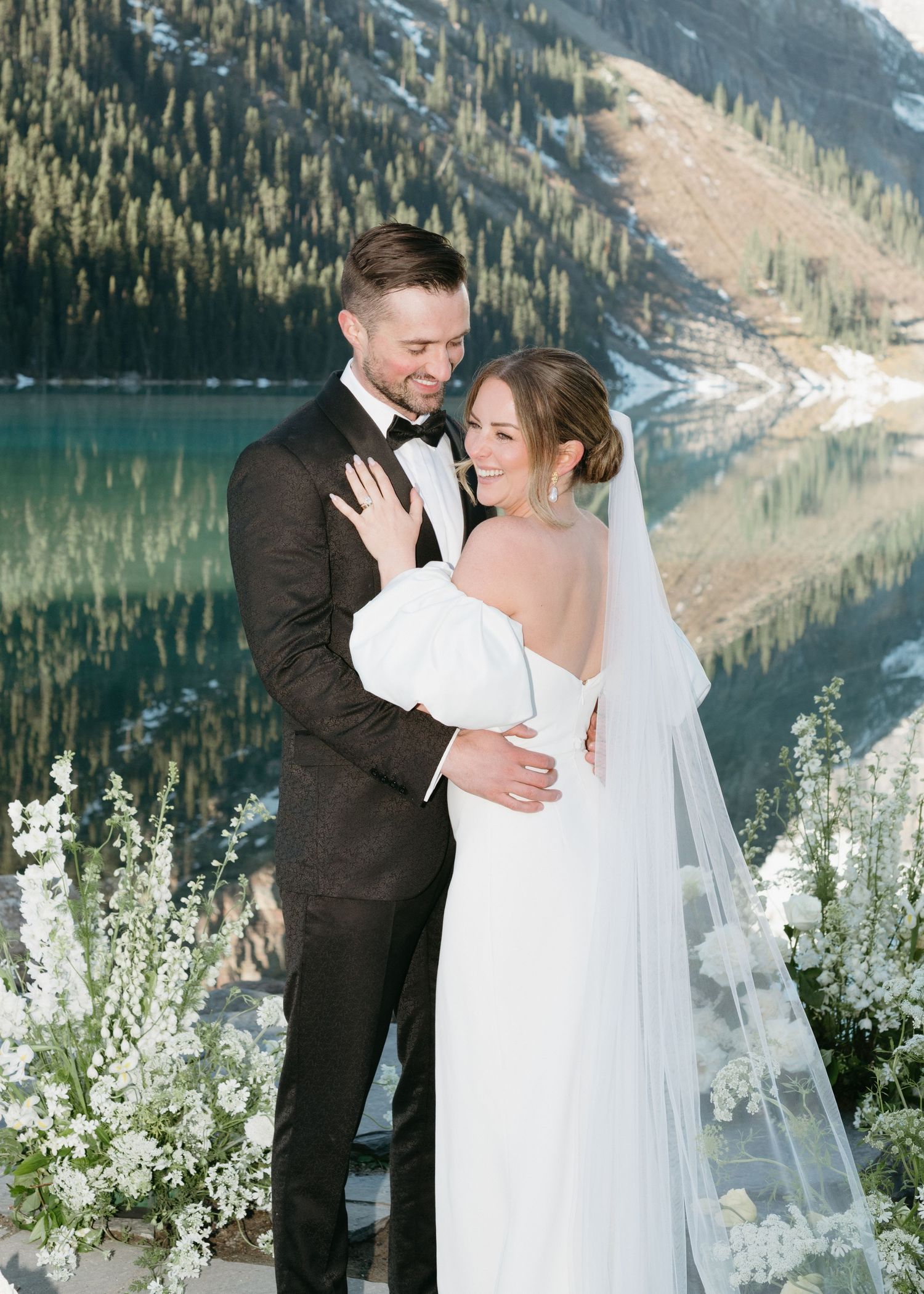 A romantic wedding photo shoot against a stunning mountain lake backdrop with evergreen trees and snowy peaks.