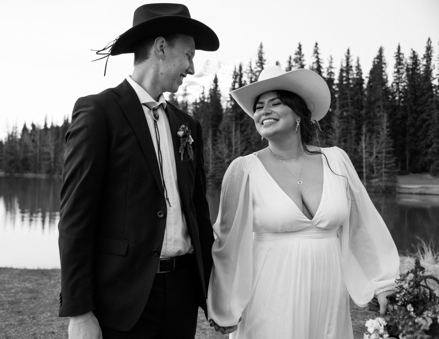 Black and white photo of a couple in western attire sharing a joyful moment by a lake. Banff and Calgary Wedding Photographer