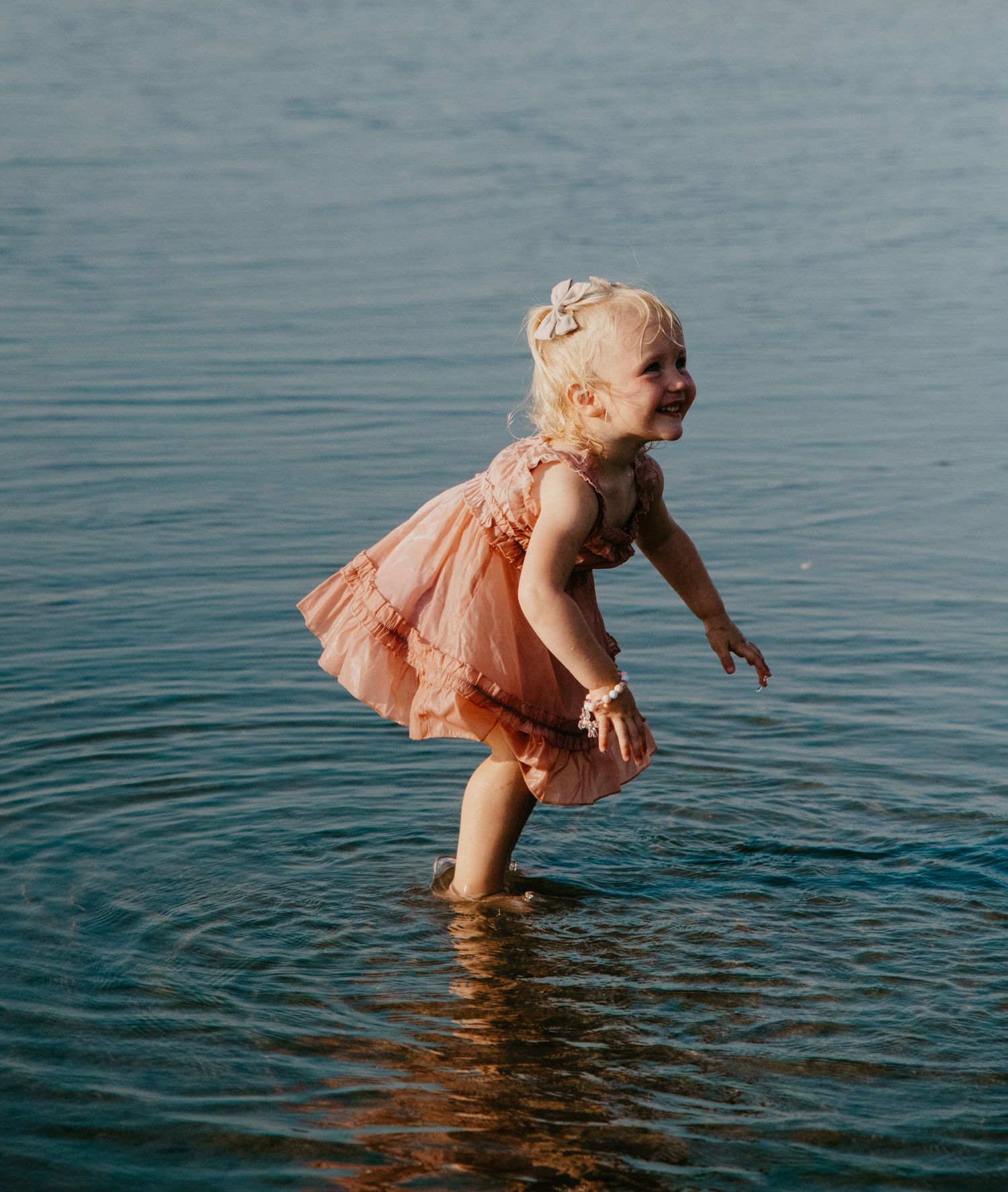 A small child in a peach dress splashes joyfully in turquoise water during a sunny day. Calgary Alberta Family Photographer.