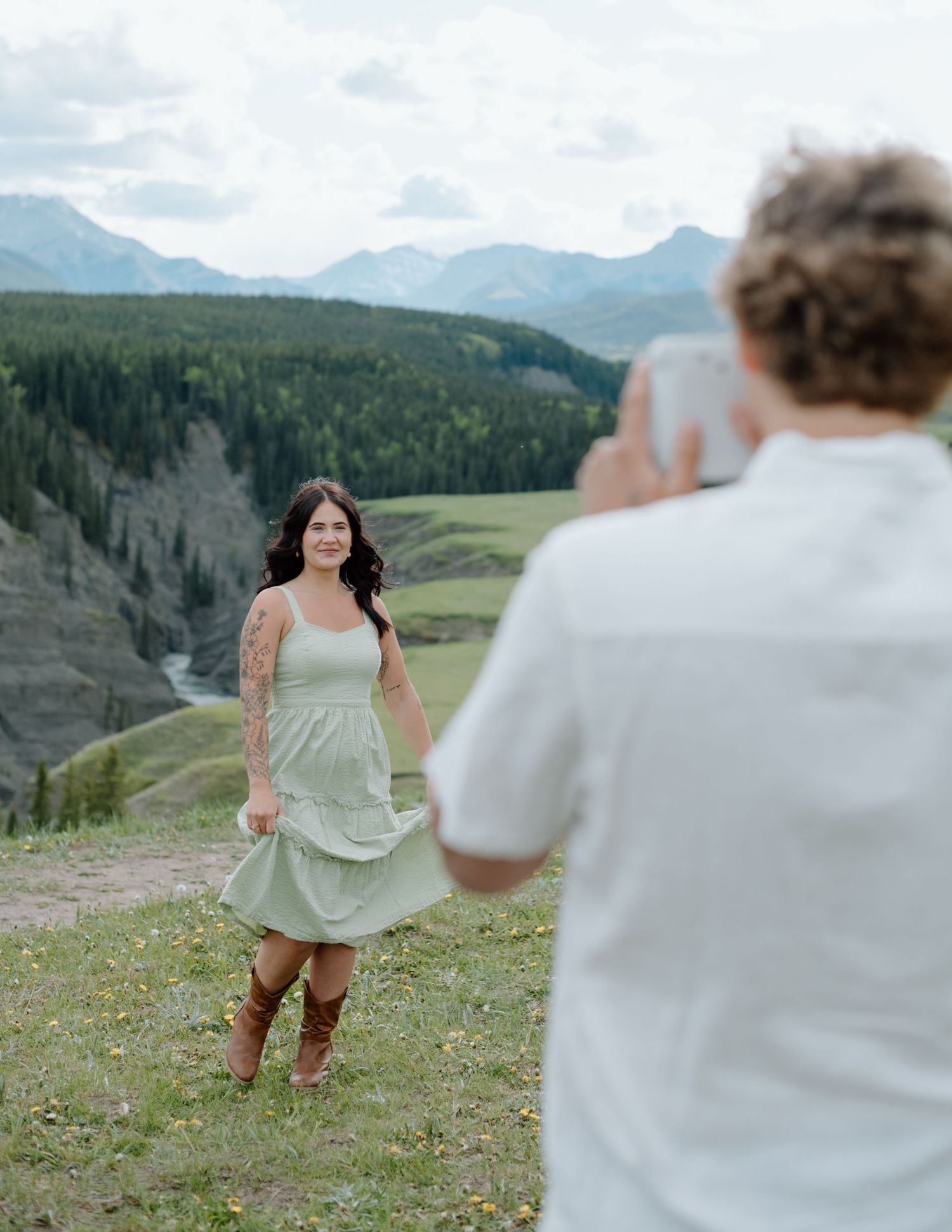 A couple taking photos in a scenic mountain meadow with rolling hills in the distance in Calgary, Alberta Calgary Lifestyle and Engagement