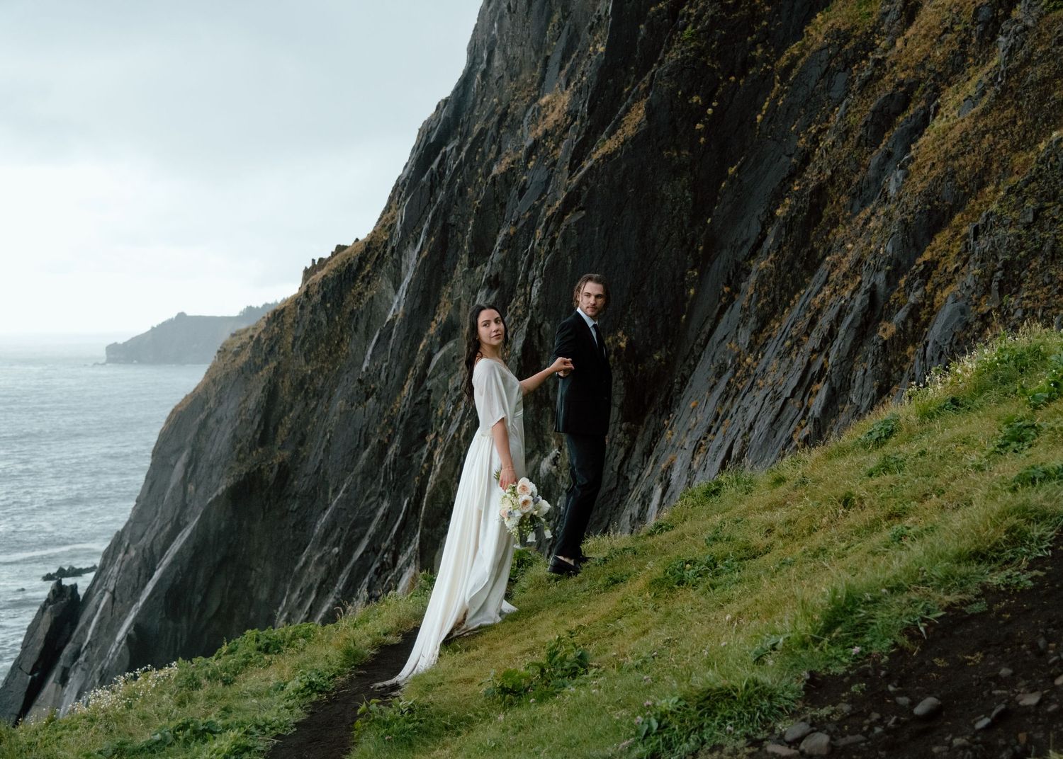 Wedding couple stands against rugged cliffs with ocean waves crashing in the background. Calgary Wedding Photographer