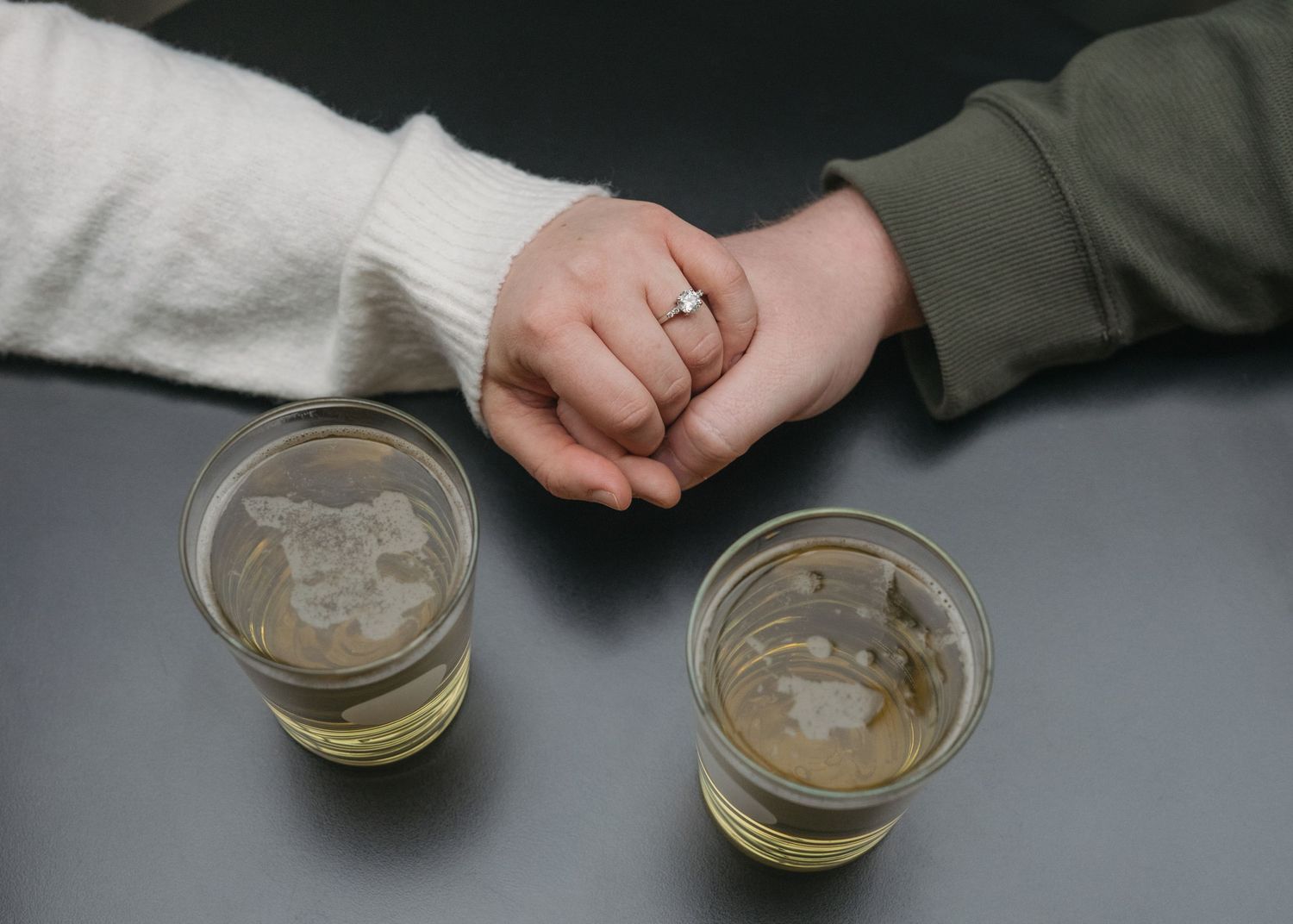 Two hands reach across a table with beer glasses, one wearing a gold ring, sharing an intimate moment over drinks.
