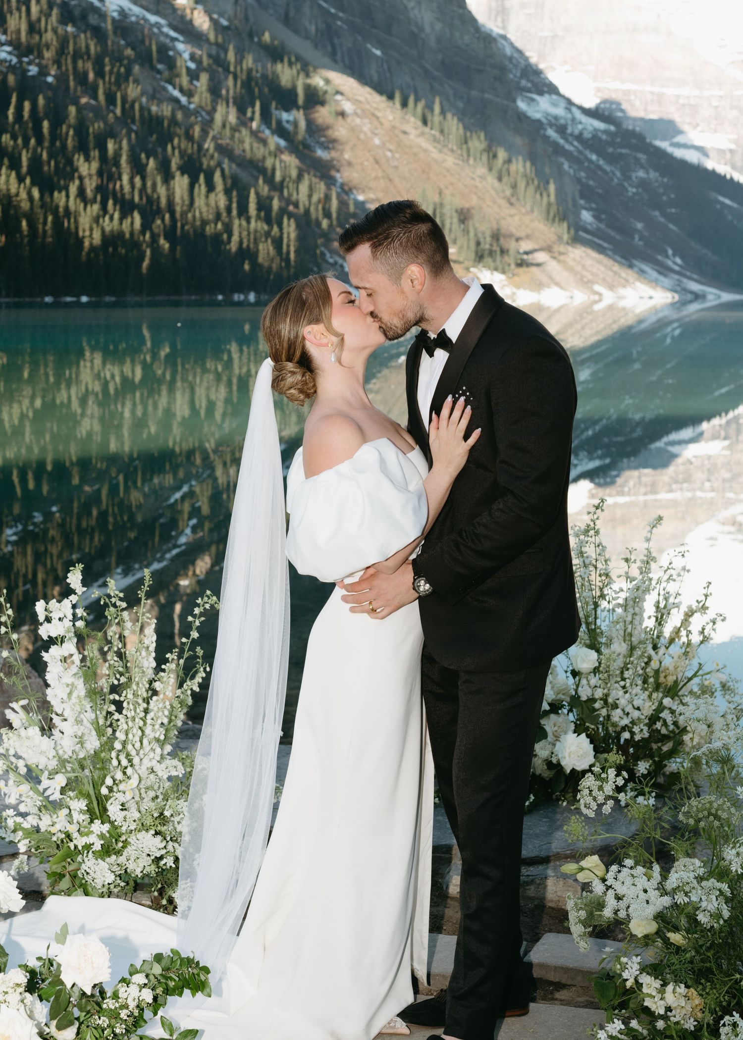 A wedding couple embraces near a pristine mountain lake surrounded by white floral arrangements and snow-capped peaks.