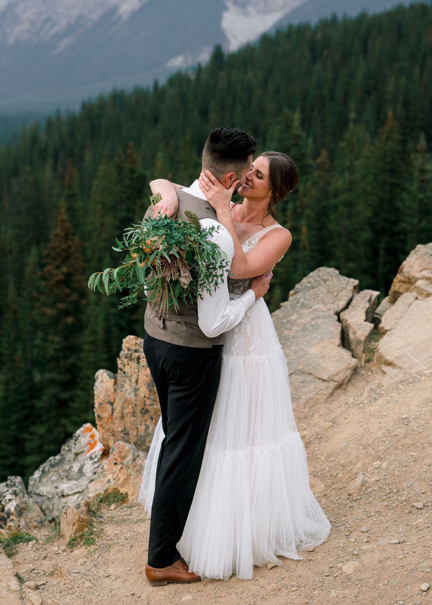A couple shares a loving embrace on a mountain ridge overlooking evergreen forests in majestic golden hour lighting.