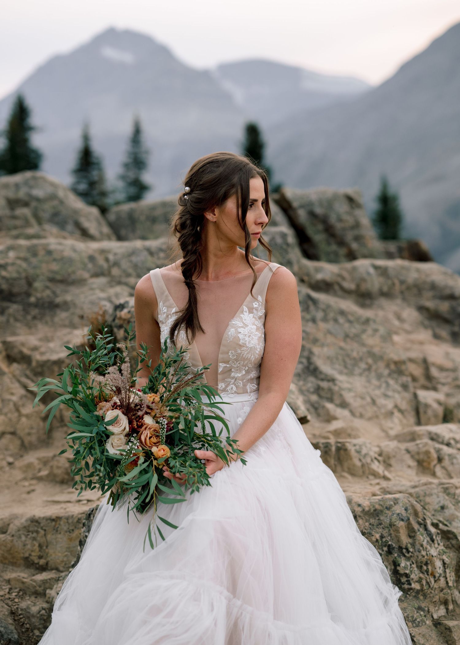A bride in a flowing white gown holds a rustic bouquet against a mountainous backdrop.