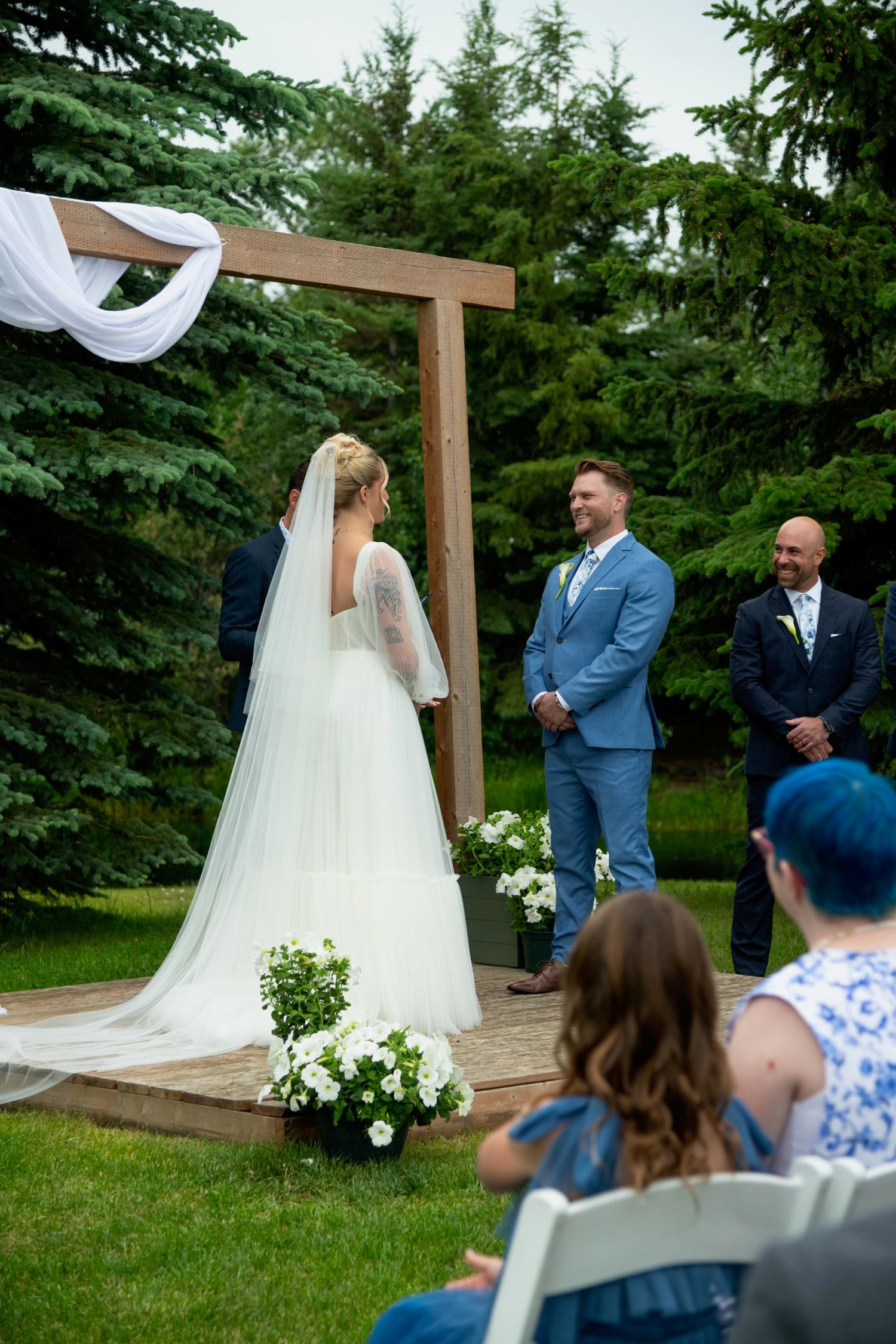 An outdoor wedding ceremony under a wooden arch surrounded by evergreen trees. Calgary Wedding Photographer