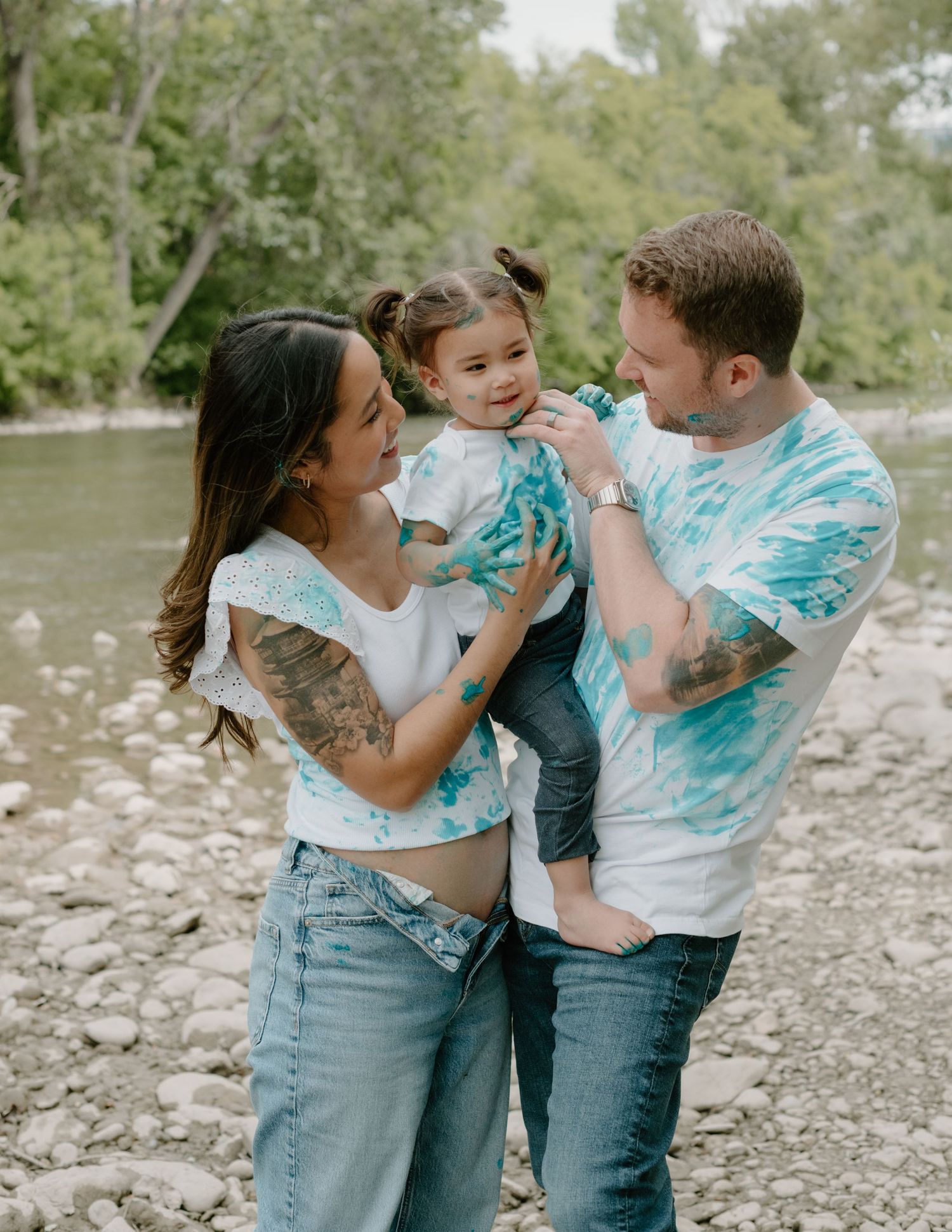 A family wearing tie-dye shirts shares a joyful moment together by a riverside on a summer day. Calgary Alberta Maternity Photographer