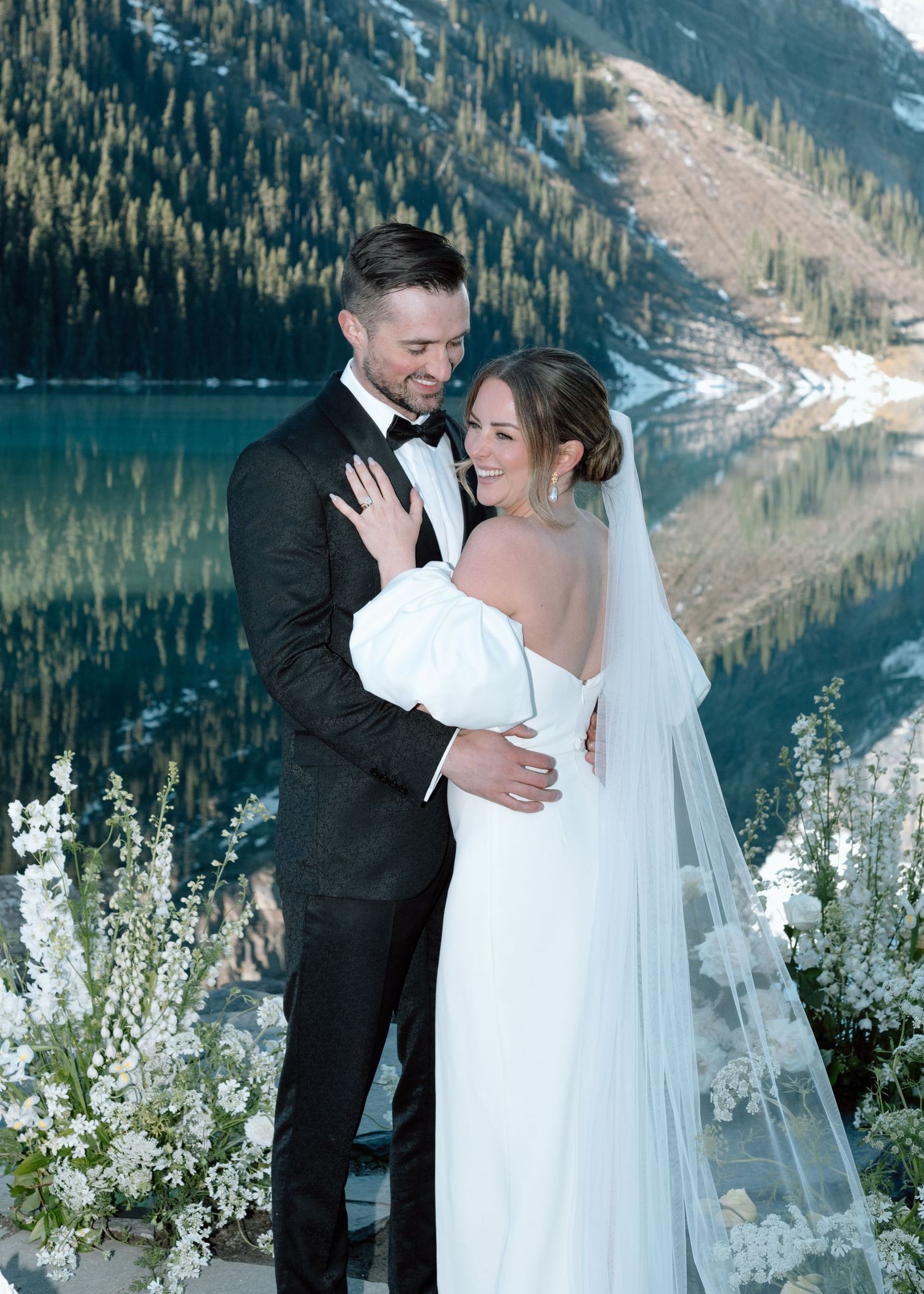 Romantic wedding couple embracing by mountain lake with floral decorations at Lake Louise, Alberta. Calgary Wedding Photographer.