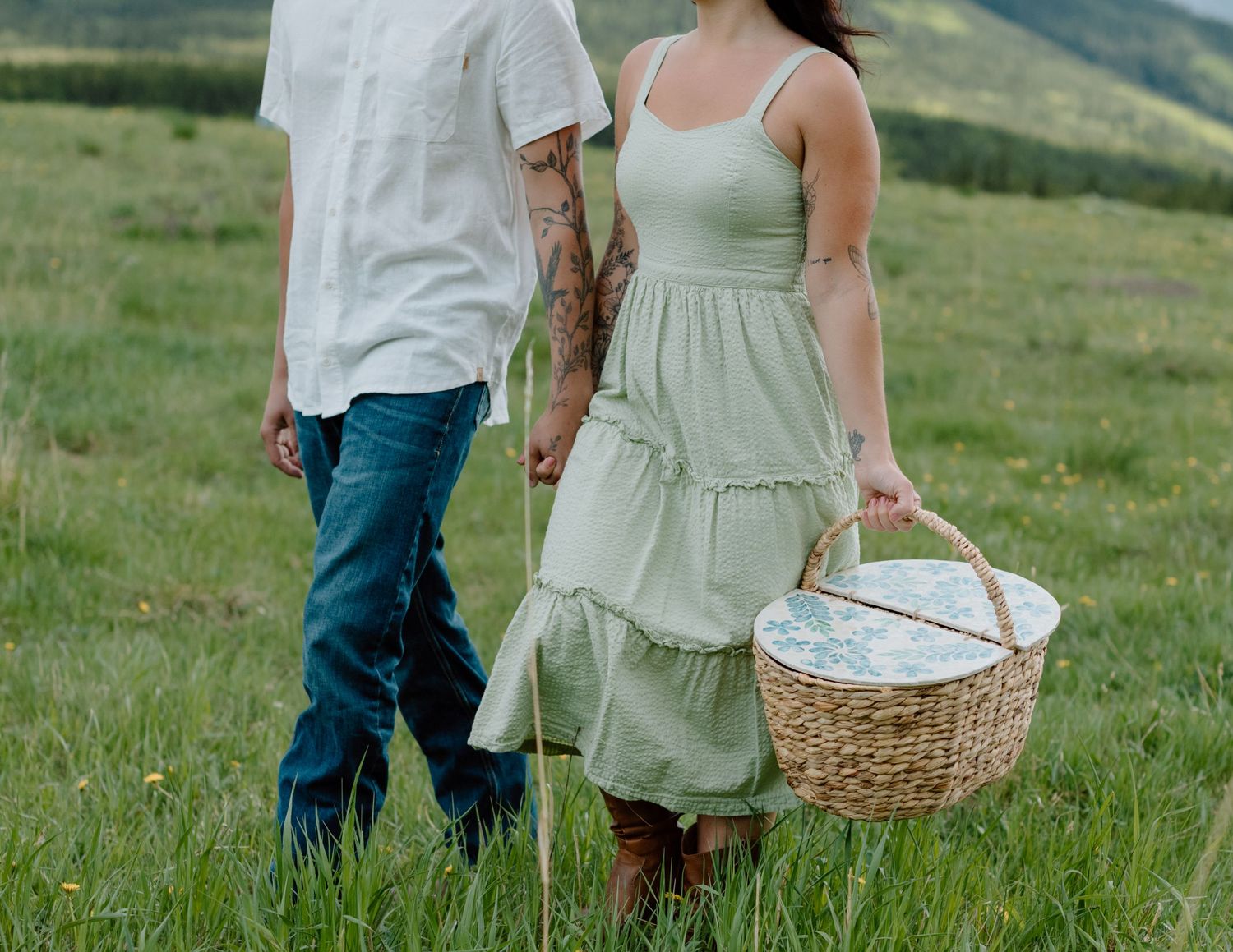 A couple walks through a mountain meadow with a picnic basket at sunset. Calgary Alberta Engagement Photographer.