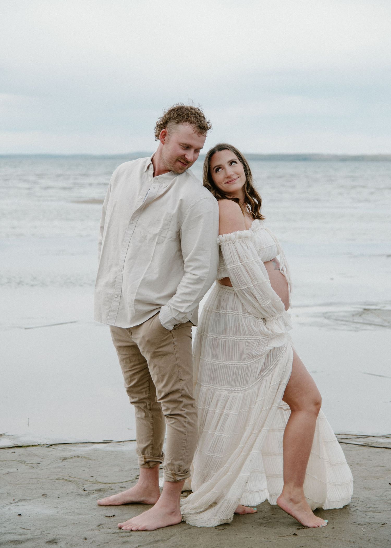 Couple poses intimately on sandy beach wearing light coloured clothing in Sylvan Lake. Calgary Maternity Photographer.