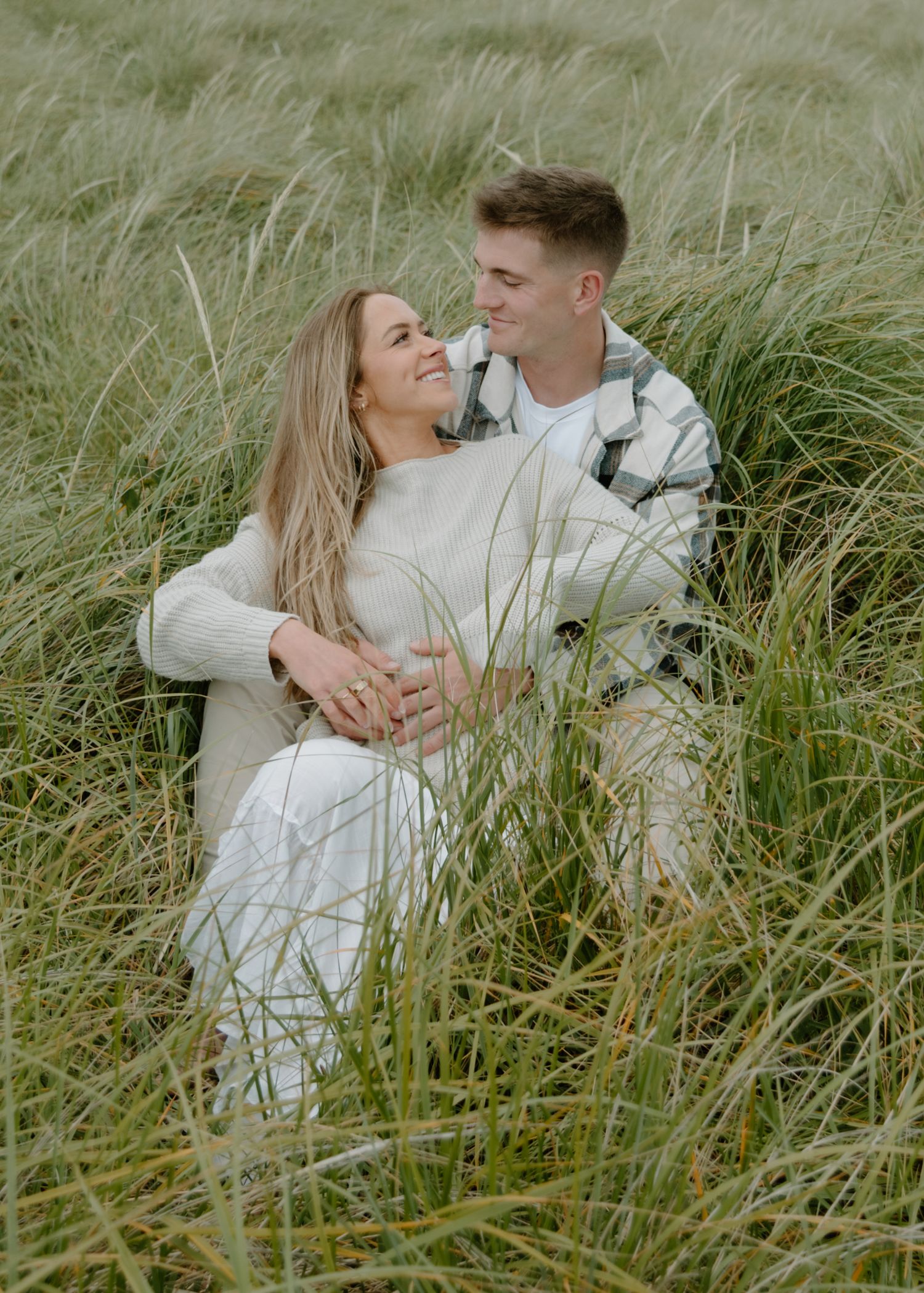 Couple in white clothing embraces among tall swaying grass on Oregon Coast. Calgary Lifestyle and Engagement Photographer.