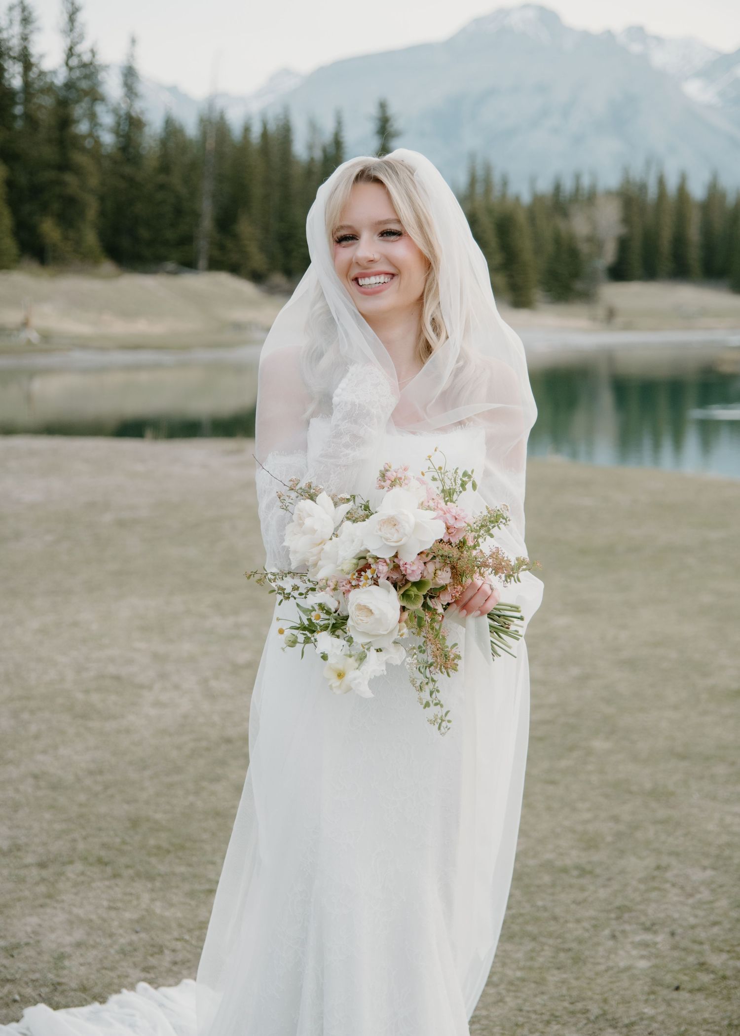 Bride in simple white gown holds delicate bouquet against mountain landscape backdrop. Banff and Calgary Wedding Photographer