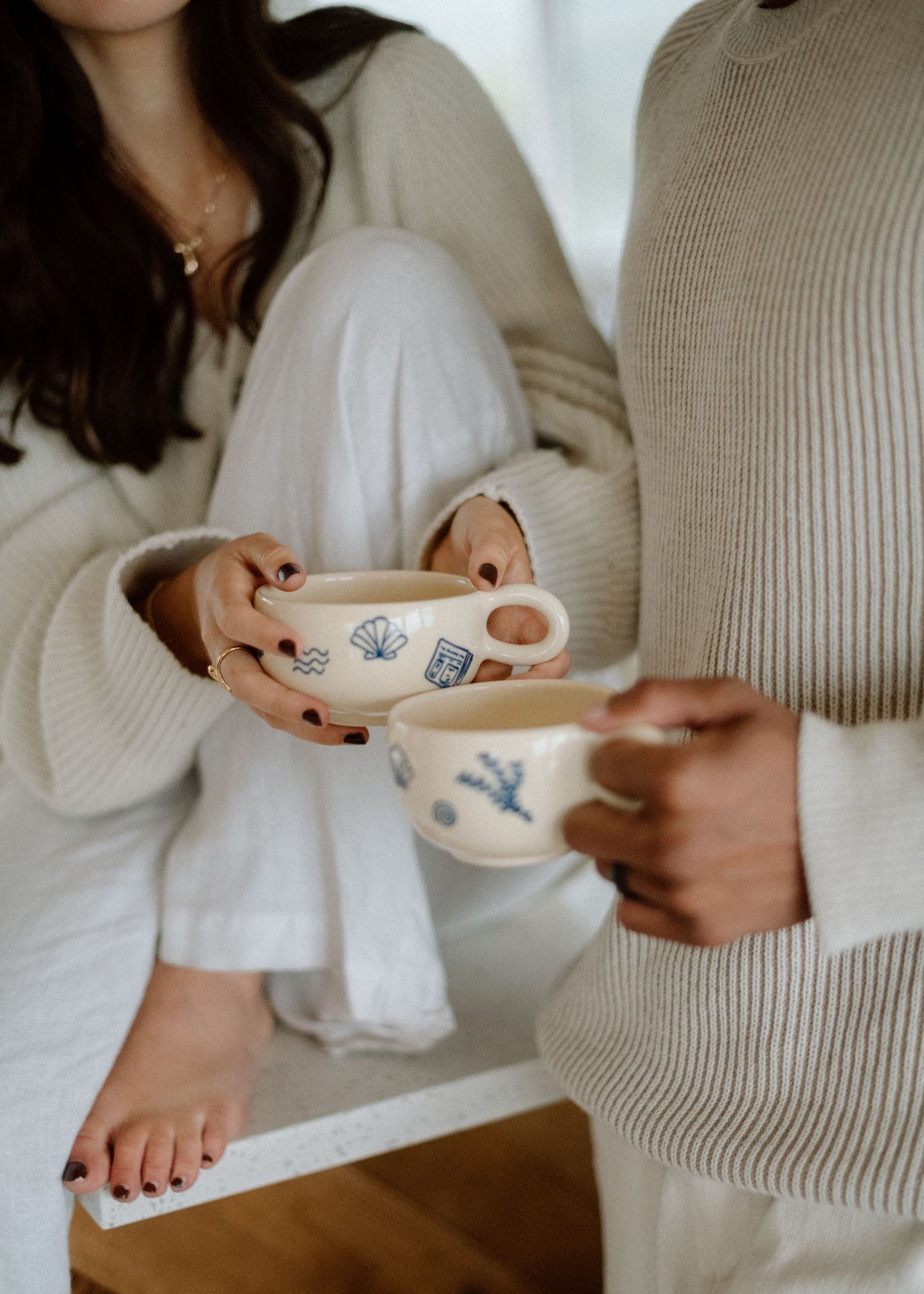 Hands holding delicate white ceramic teacups against a neutral background with warm morning light.