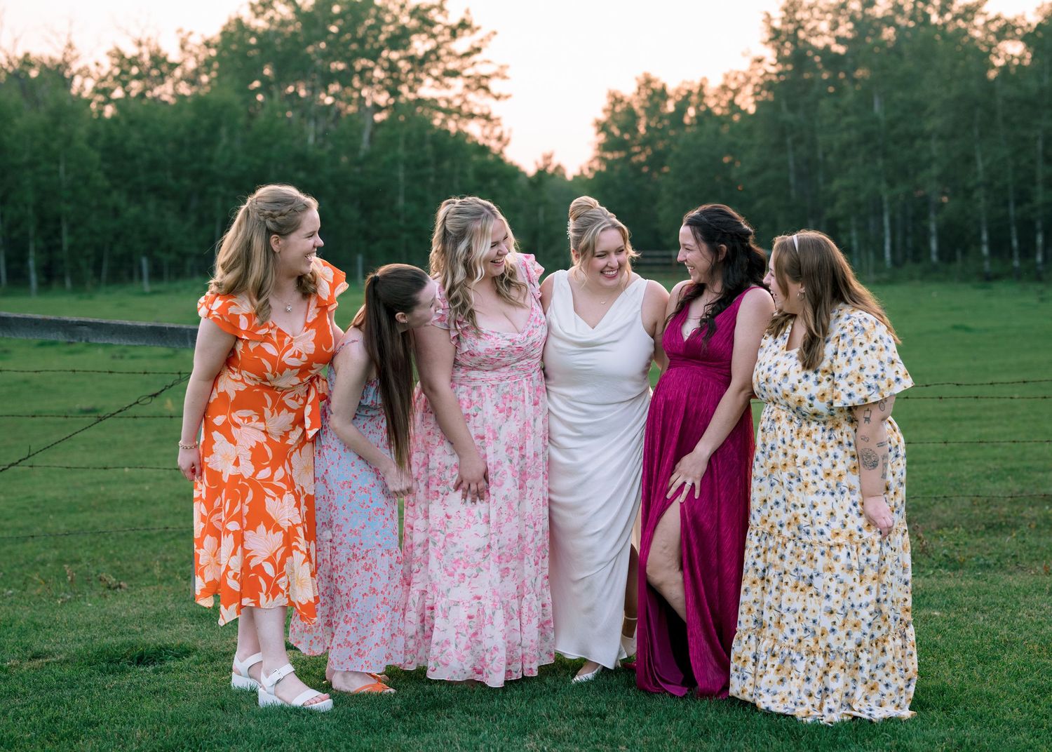A group in colorful dresses laugh together while sitting in a grassy field at sunset.