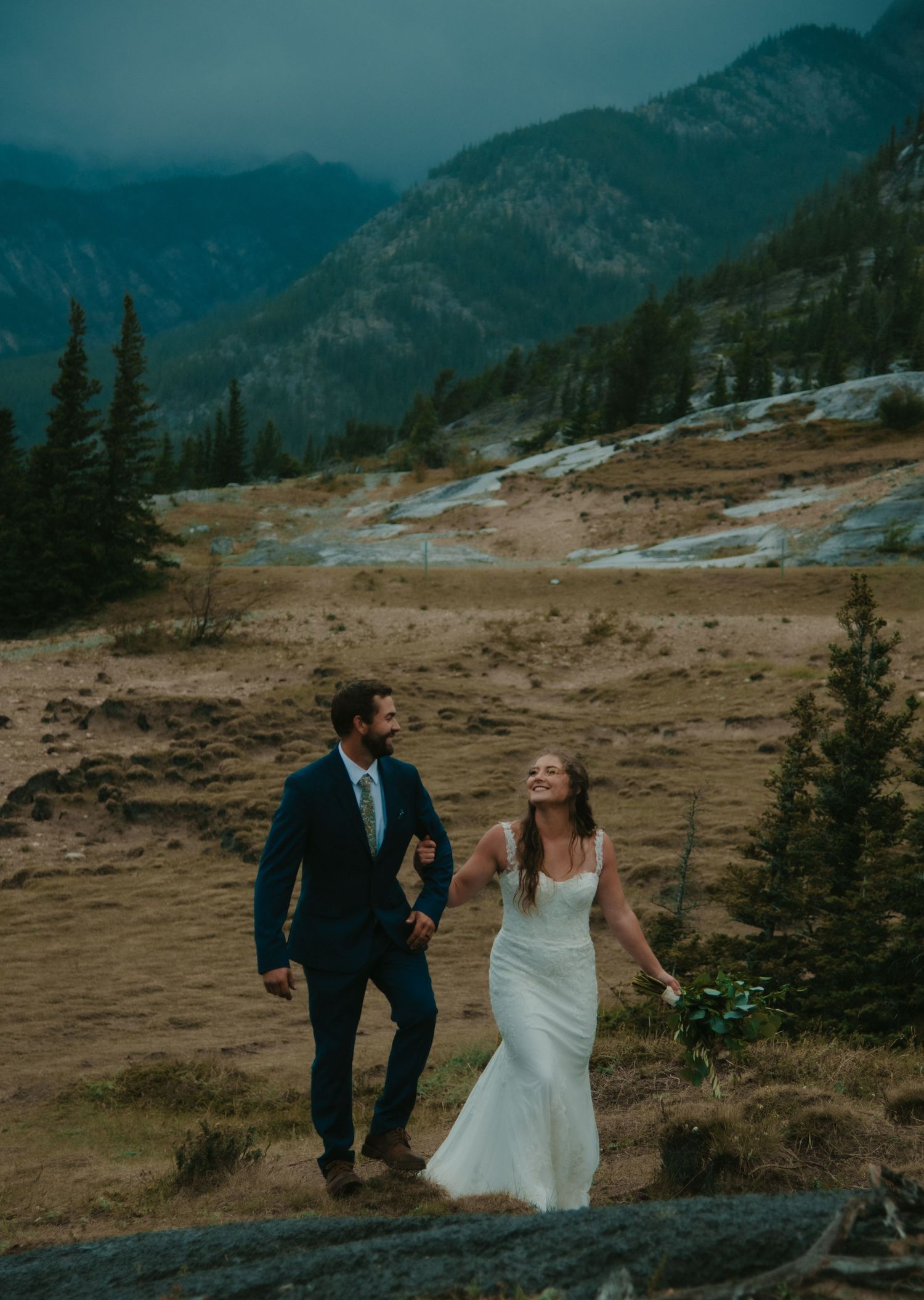 A couple walks hand in hand on a mountainside trail in wedding attire. Calgary Wedding Photographer