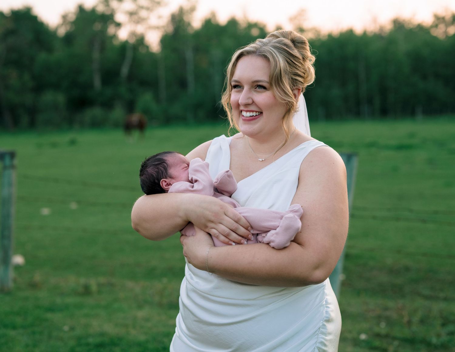 Someone in a white dress holds and plays with a baby outdoors in a green field during golden hour.