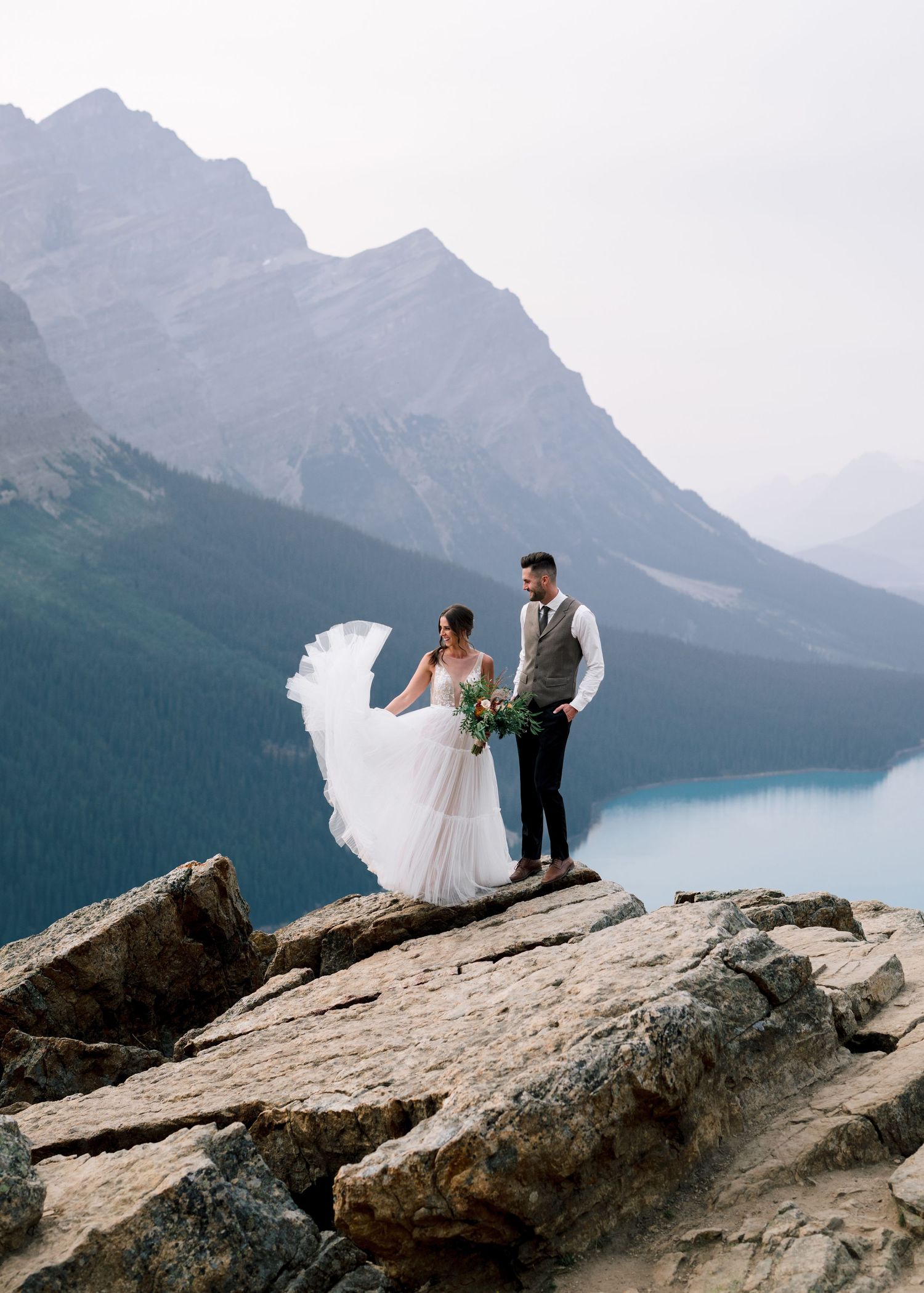 A couple in wedding attire stands on a rocky cliff overlooking a stunning mountain lake in the Canadian Rockies.