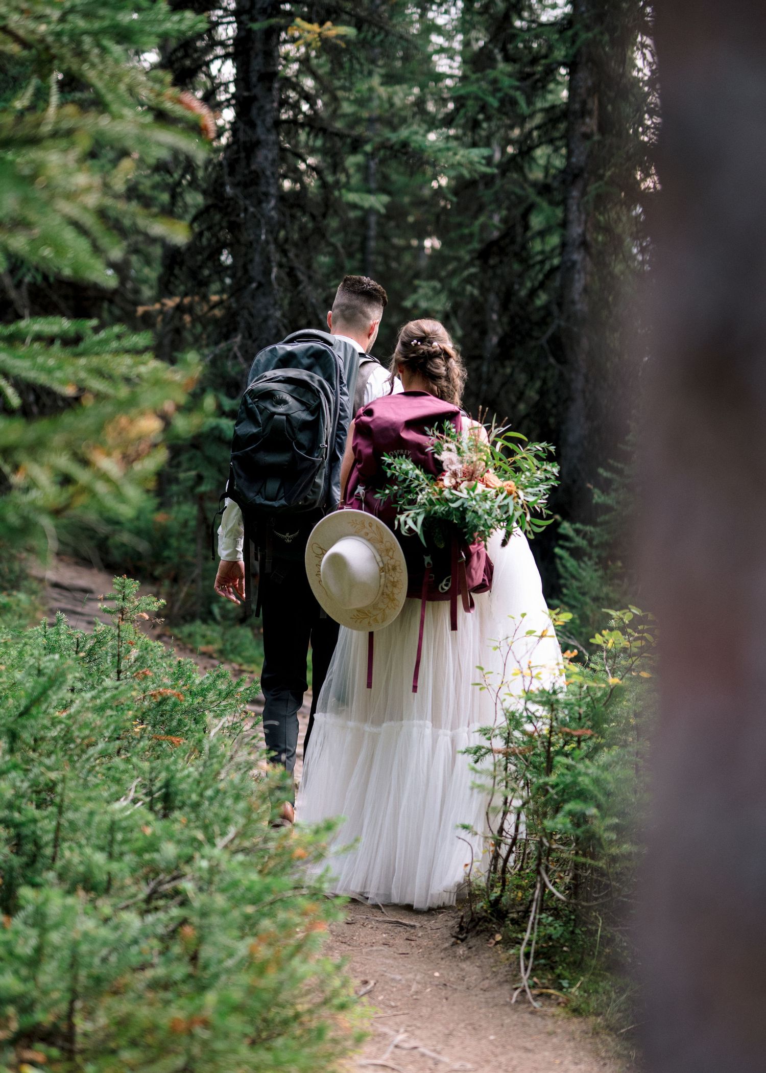 A person in a white dress and someone in dark attire walk along a wooded forest trail during an outdoor ceremony.