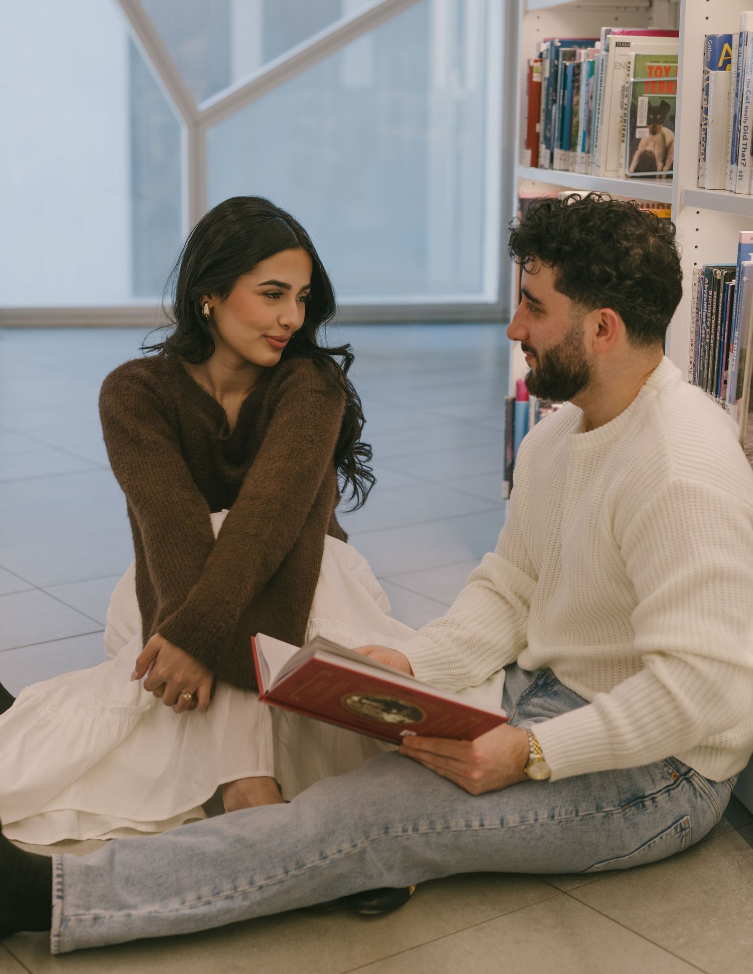 Couple lying on floor reading books together in a cozy indoor setting wearing contrasting brown and white outfits.