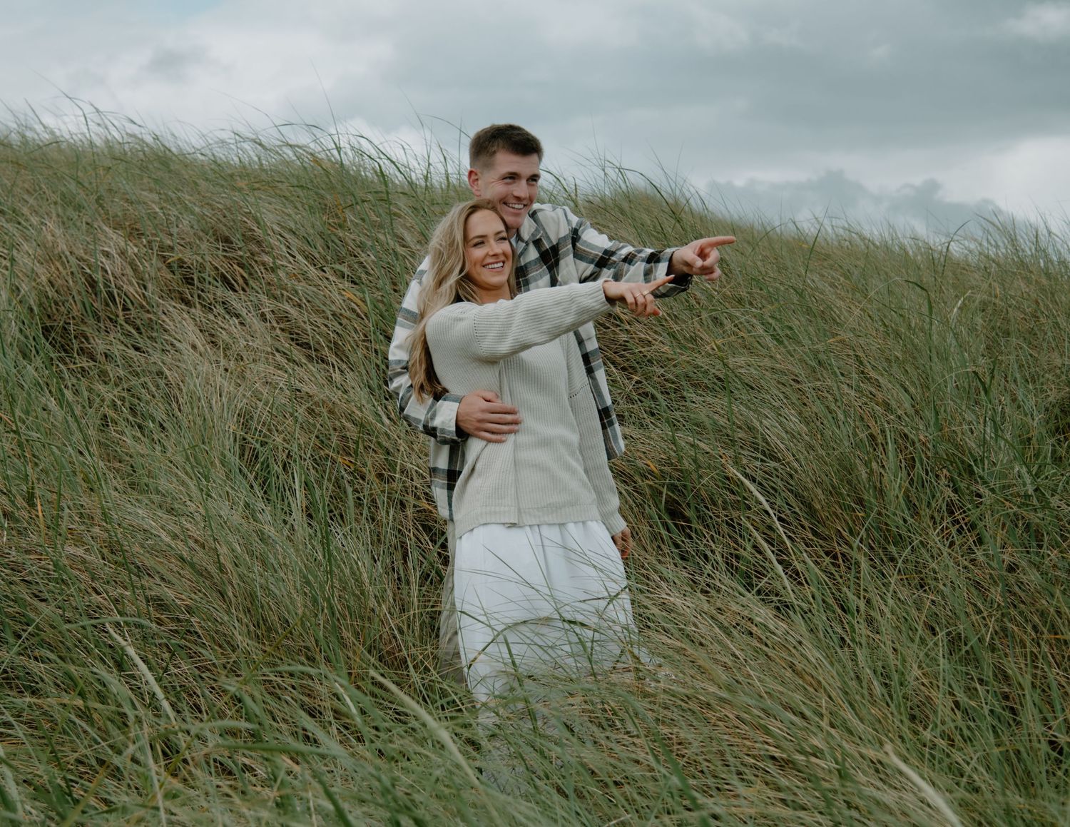 A couple shares a playful moment while pointing at something in the distance among beach grass on Oregon Coast. Calgary Lifestyle and Engage