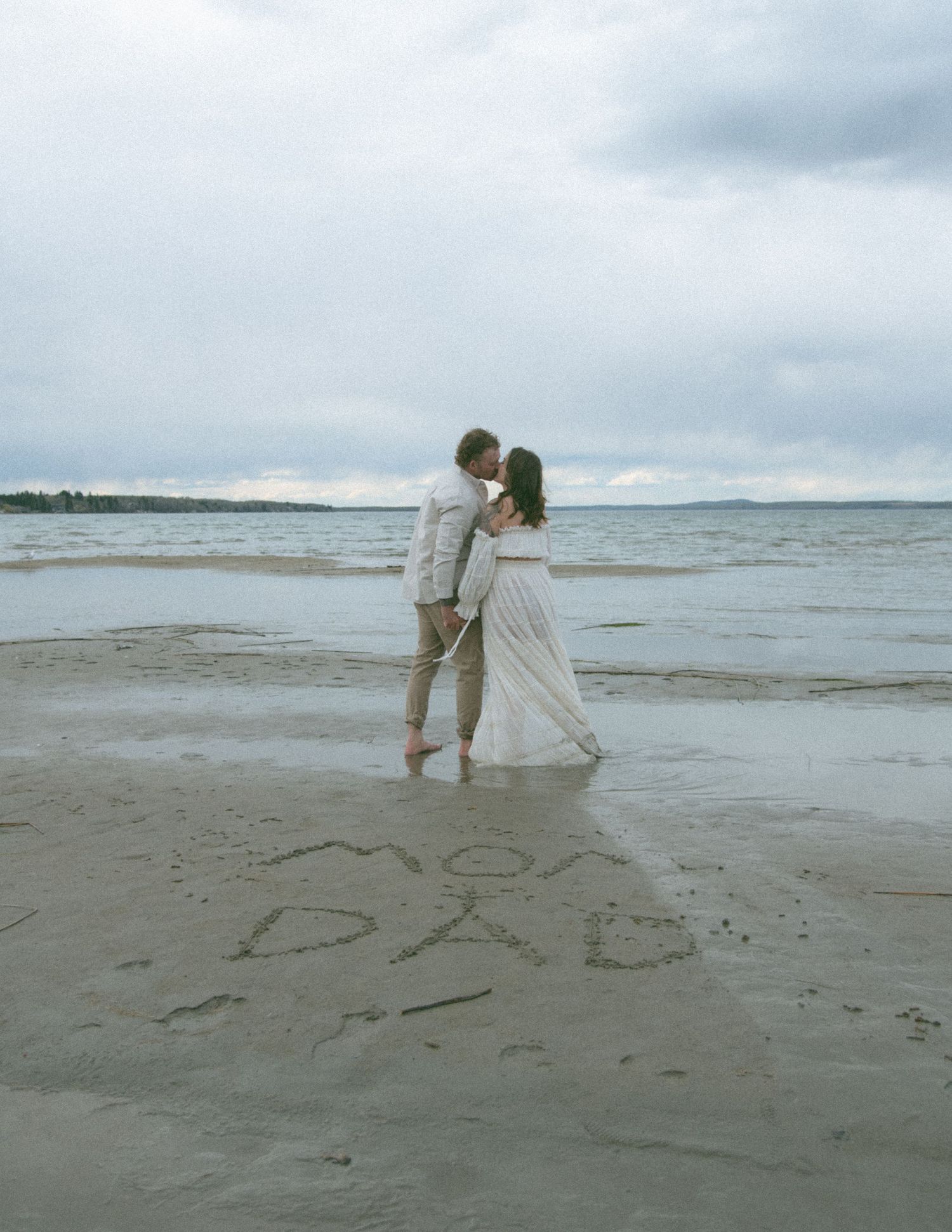 A couple in light coloured clothing standing together on a beach with cloudy skies. Calgary Alberta Maternity Photographer.