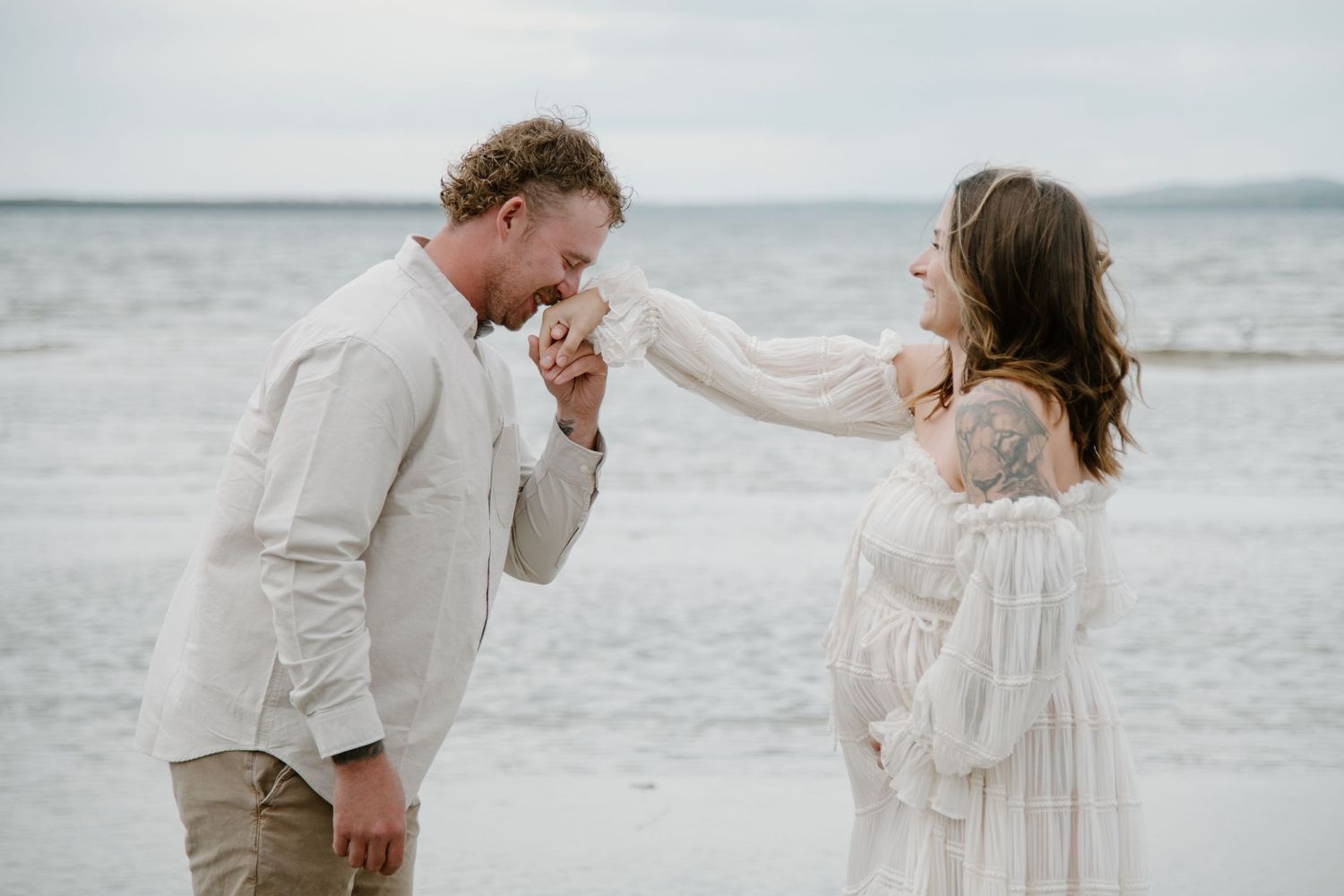 A sweet moment captured as a couple shares a romantic gesture on a cloudy beach. Calgary Alberta Maternity Photographer.