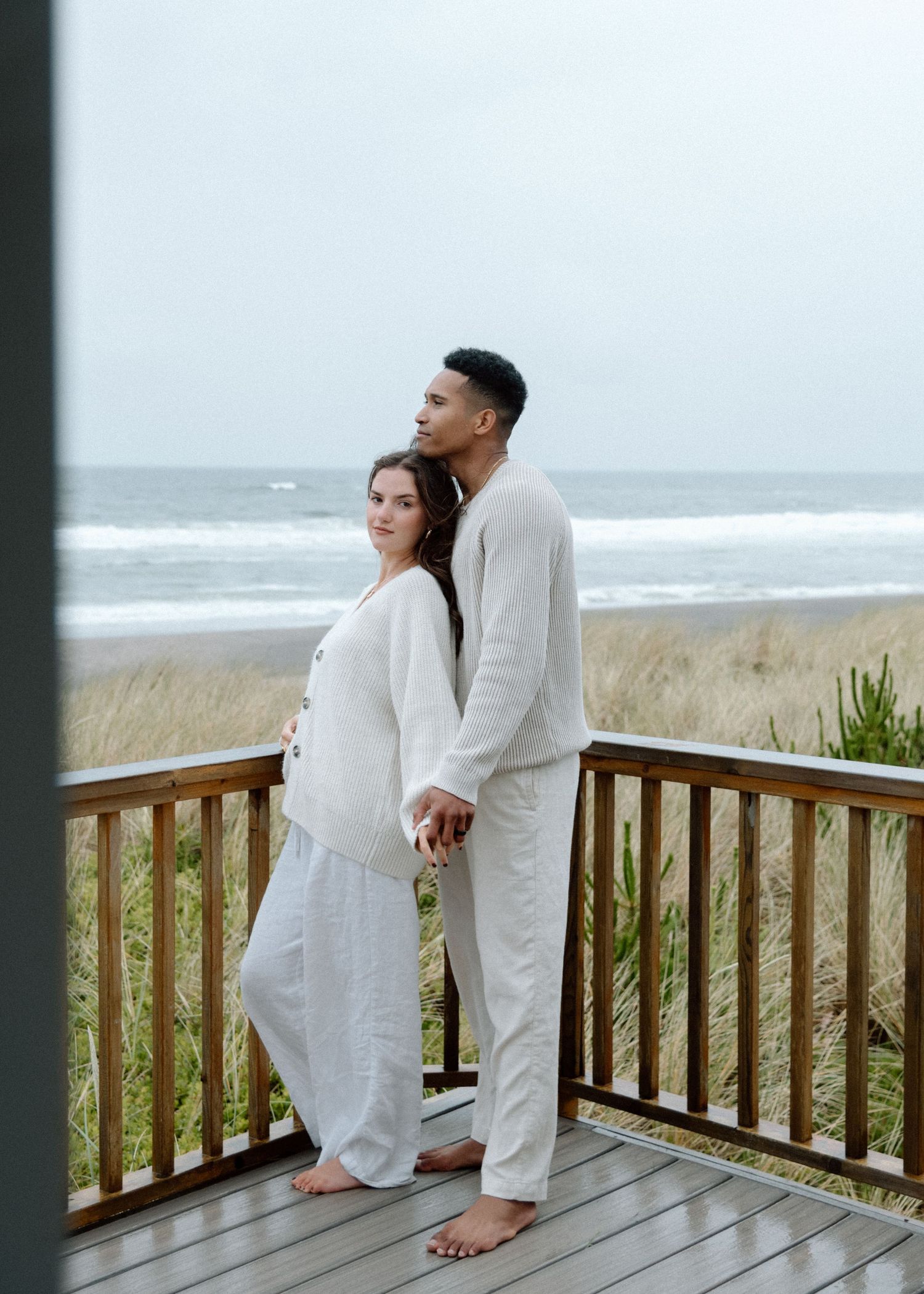 Couple in white outfits sharing tender moment on oceanfront deck with beach view.
