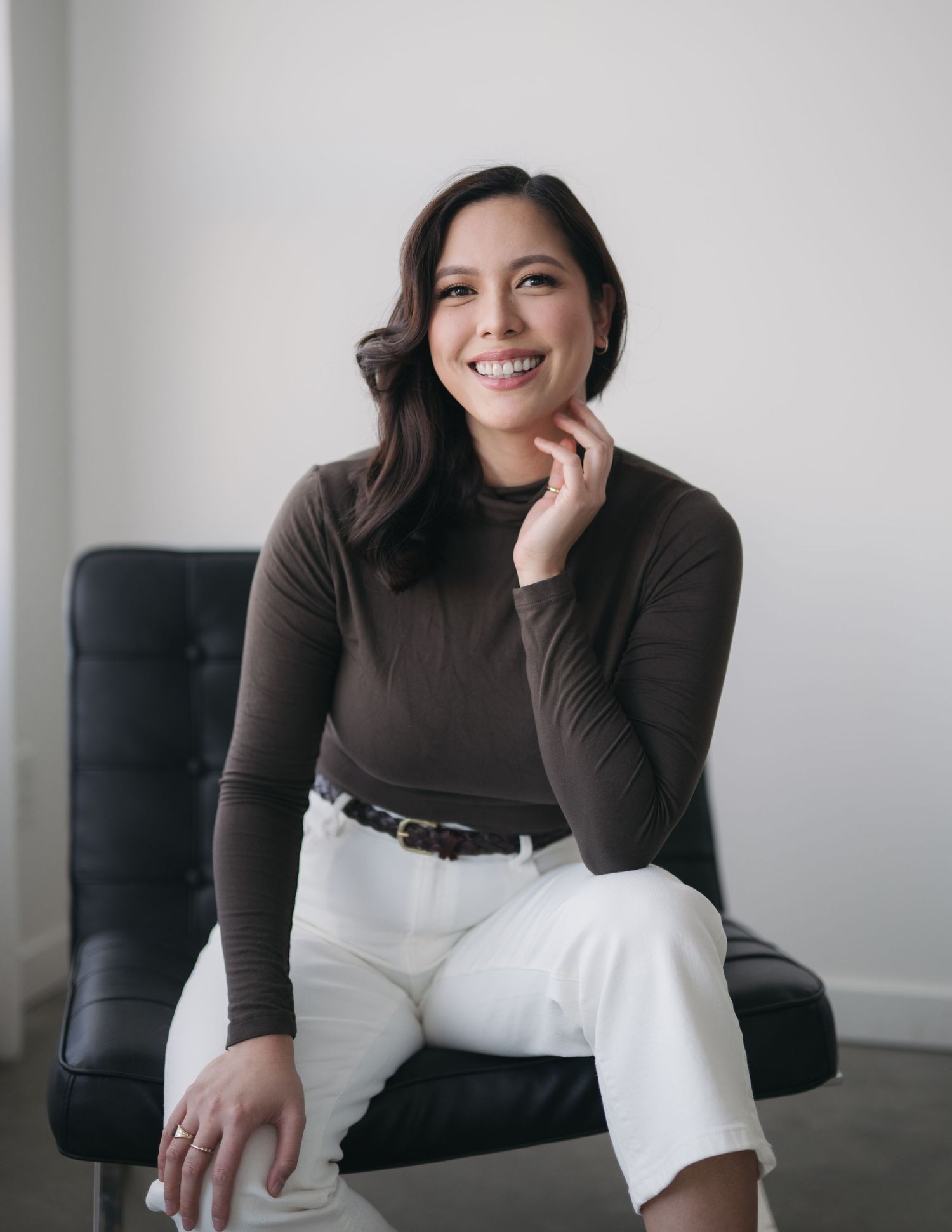 Smiling woman in brown turtleneck and white pants sits casually in dark chair with hand resting on chin.
